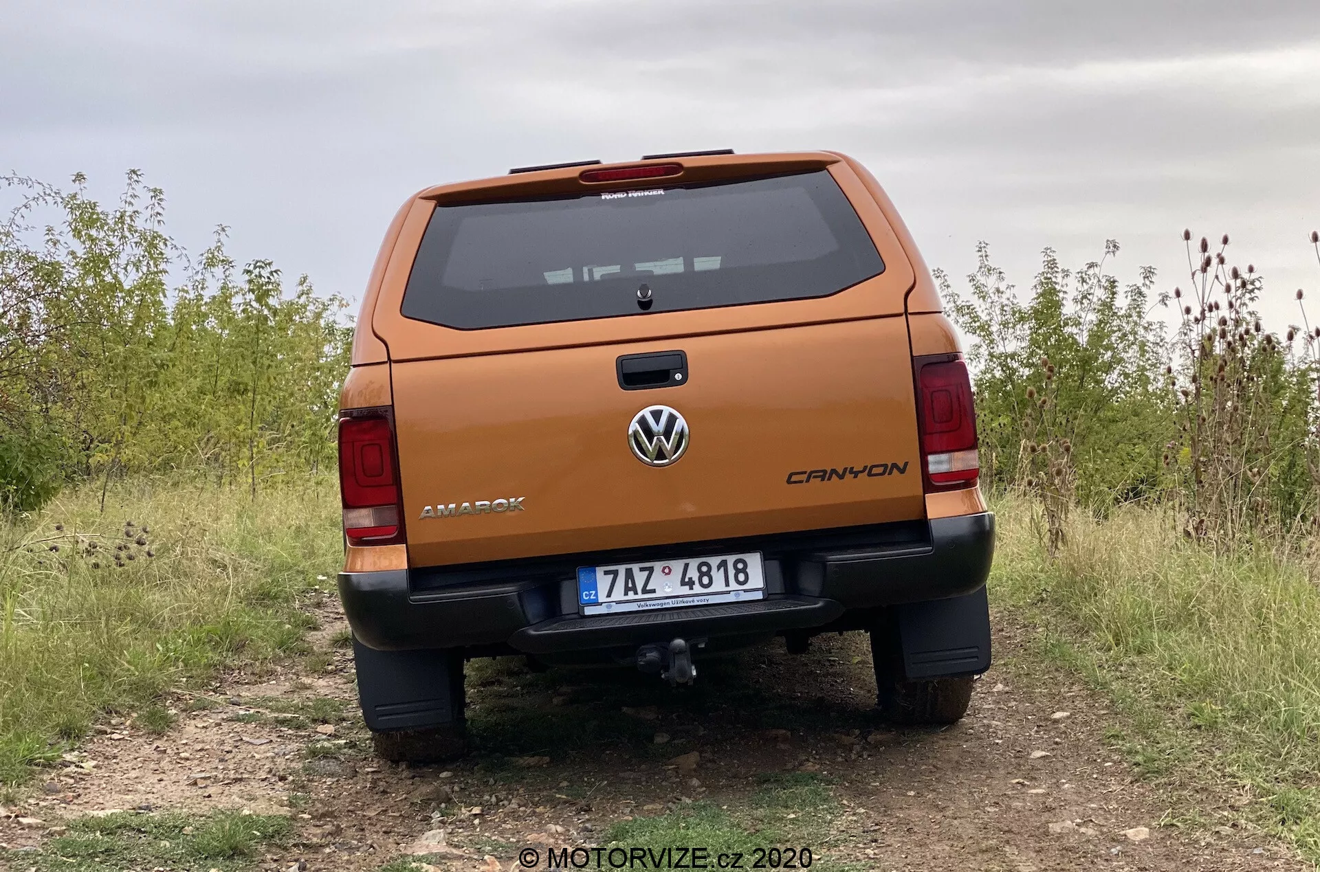 Profesionální zadní pohled na pickup Volkswagen Amarok 2016 z nízkého úhlu, zdůrazňující robustní zadní víko s nápisem 'AMAROK', logem Volkswagen uprostřed, označením výbavy 'CANYON', poznávací značkou, podrobnými zadními světly, nárazníkem s parkovacími senzory a stupátkem, tažnou tyčí, zaparkovaným na nezpevněné cestě, což naznačuje terénních schopností.
