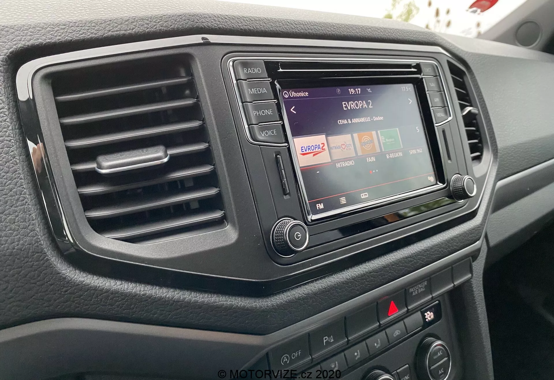 Detailní záběr na středovou konzolu VW Nutzfahrzeuge Amarok 2016 z pohledu řidiče, který ukazuje infotainment systém s rádiovou funkcí, jediný výdech klimatizace vlevo a různé ovládací tlačítka a knoflíky. Infotainment systém je zasazen do tmavého, elegantního obložení a povrch palubní desky má texturovaný povrch.