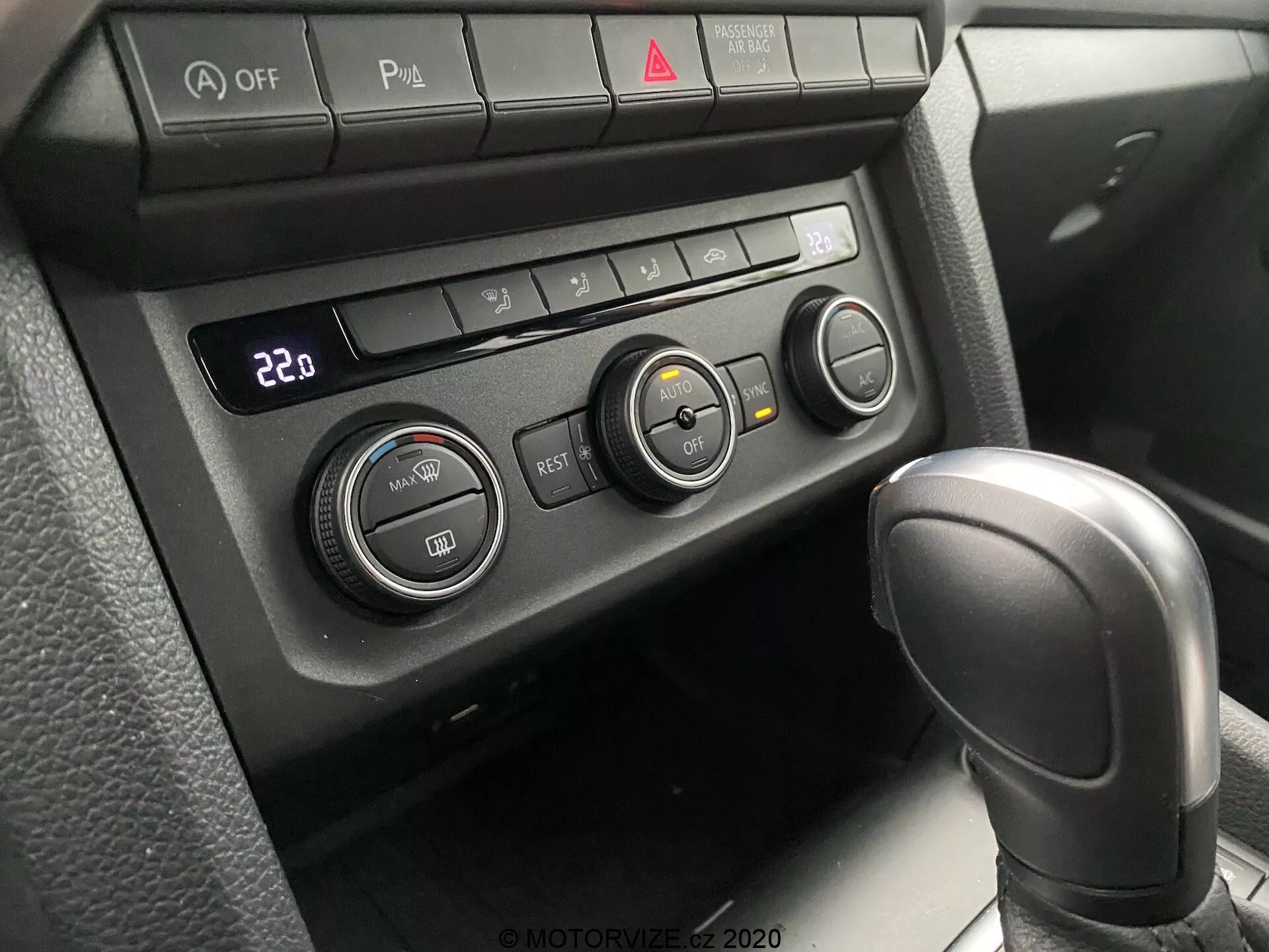Zblízka pohled na klimatizační systém a částečnou řadicí páku modelu VW Nutzfahrzeuge Amarok 2016. Perspektiva pasažéra nebo řidiče se zaměřuje dolů na středovou konzolu. Obsahuje koženou automatickou řadicí páku v poloze 'Park' a klimatizační panel nastavený na 22°C se třemi otočnými ovladači a několika funkčními tlačítky. Tlačítka zahrnují vypnutí Auto Stop/Start, přepínač parkovacích senzorů, vyhřívaný volant a vyhřívané sedadlo spolu s červeným tlačítkem výstražných světel uprostřed. Měkké a rozptýlené osvětlení naznačuje dobře zastíněný interiér.