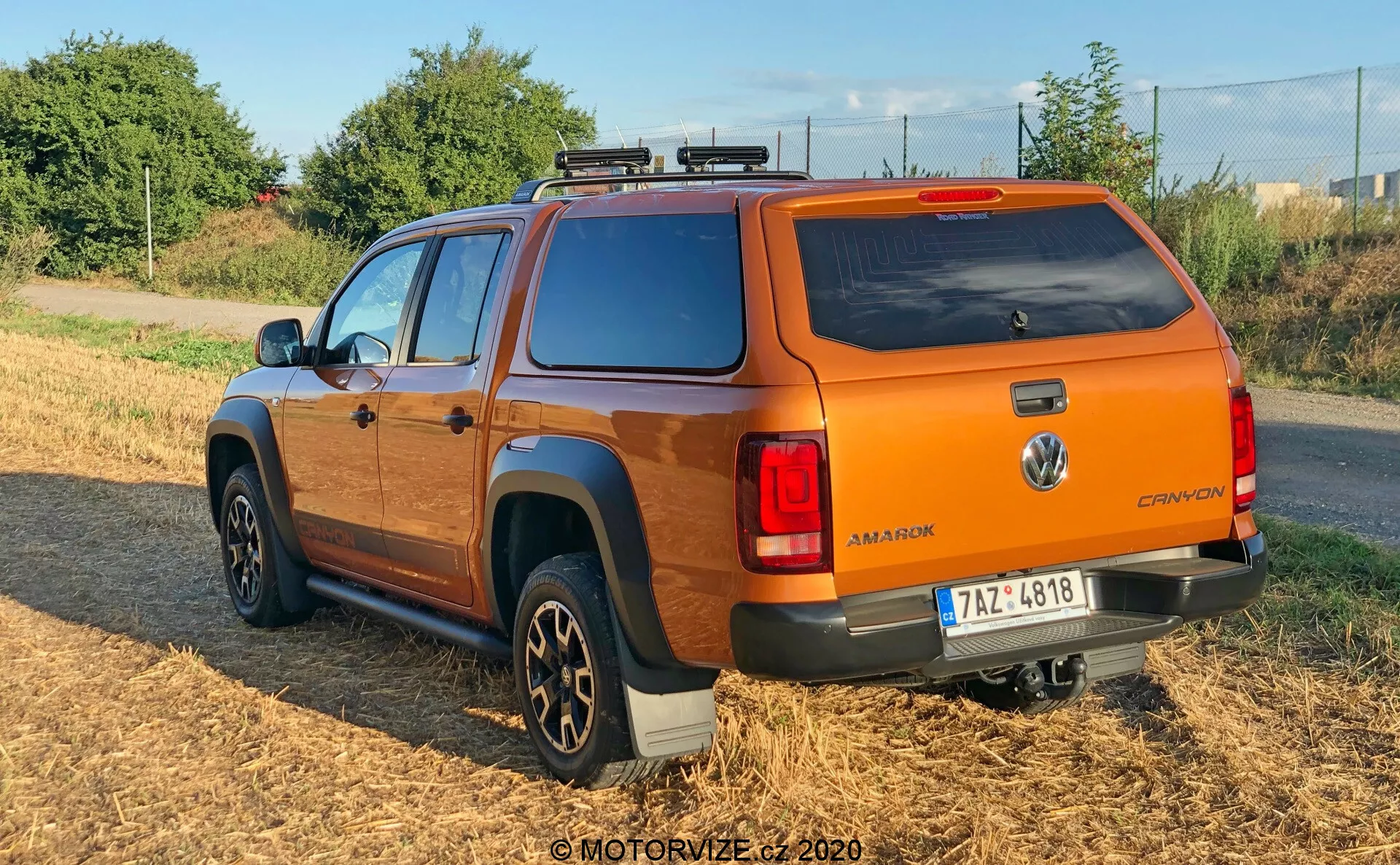 Tříčtvrteční zadní pohled na oranžový pickup Volkswagen Amarok 2016 s pevnou střechou, střešním nosičem a vícepaprskovými slitinovými ráfky. Vozidlo je zaparkováno na travnaté ploše s mírným přírodním osvětlením, zobrazuje odznak Volkswagen, název modelu 'AMAROK' a označení 'CANYON' a moderní zadní světla. Tmavě zbarvené boční stupátky a konfigurace dvojité kabiny zvýrazňují jeho užitkovou a cestovní funkčnost.