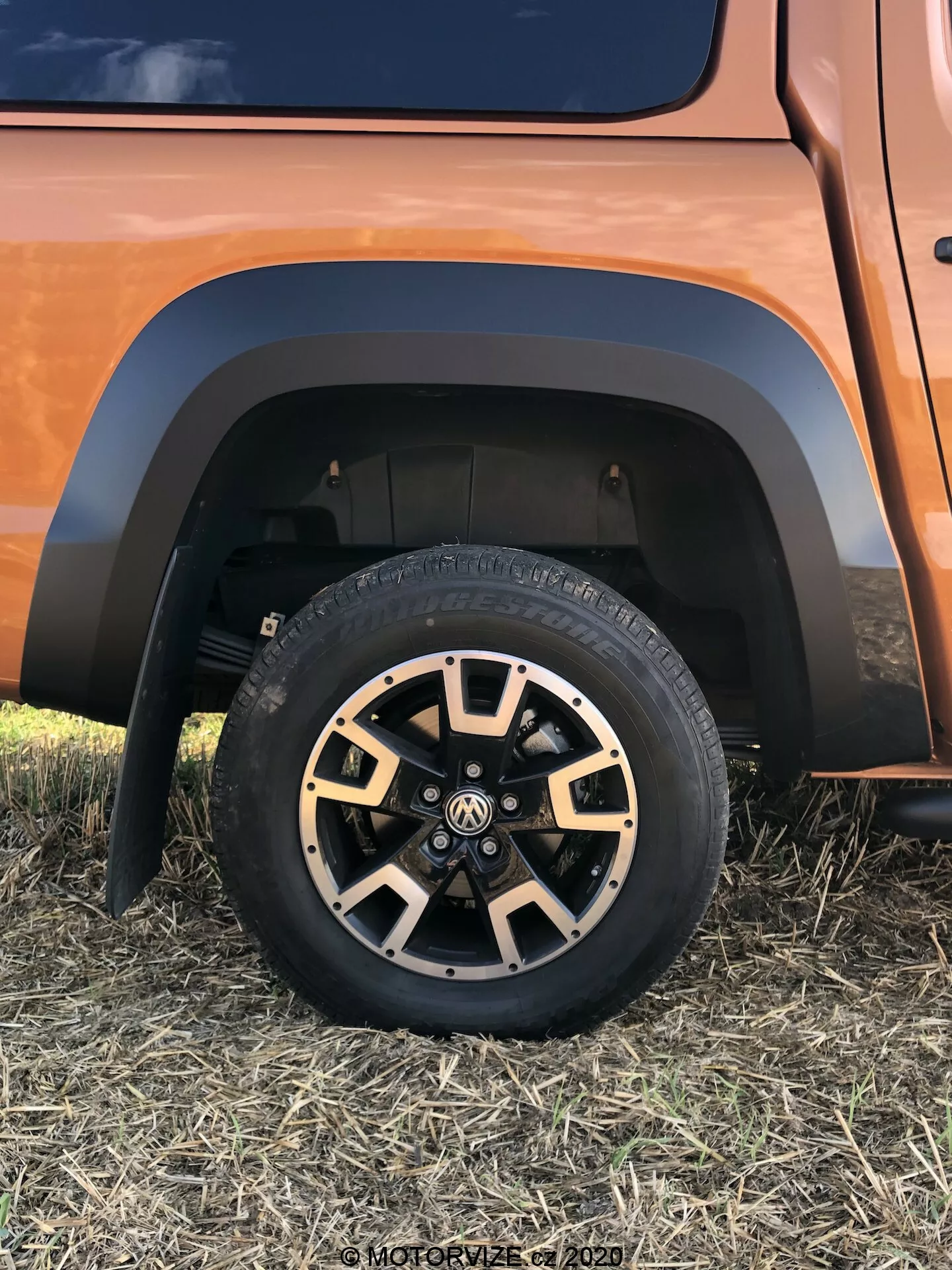 Detailní pohled na spodní část vozu VW Nutzfahrzeuge Amarok 2016, zobrazující robustní terénní pneumatiku na vícepaprskovém slitinovém kole s černými a kovovými povrchovými úpravami, kontrastní blatník a část odpružení v zemitě nebo měděně zbarveném voze, pořízený z nízkého předstranového úhlu.