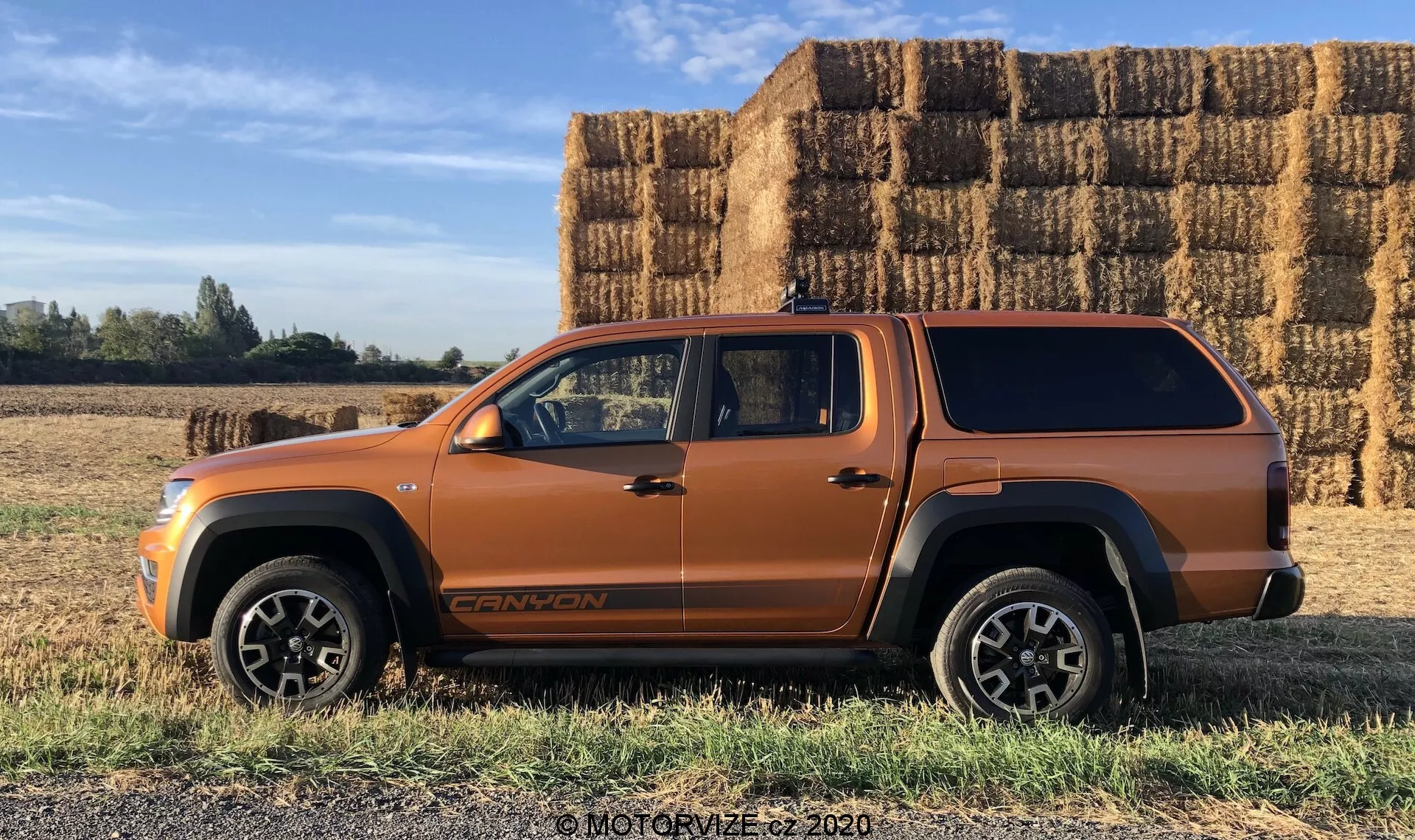 Boční profil kovově oranžového SUV-typu vozu, pravděpodobně středně velkého SUV nebo pickup s prodlouženou kabinou a krytem ložné plochy, zaparkovaný na plochém travnatém povrchu s velkými obdélníkovými balíky sena v pozadí za slunečného, jasného dne. Vozidlo ukazuje výrazné linie karoserie, velké oblouky kol, černé dolní lišty karoserie, robustní hliníková kola, viditelný znak nízko na předních dveřích a střešní anténu. Fotografie zvýrazňuje potenciální terénní schopnosti a bezproblémový design krytu ložné plochy vozu.