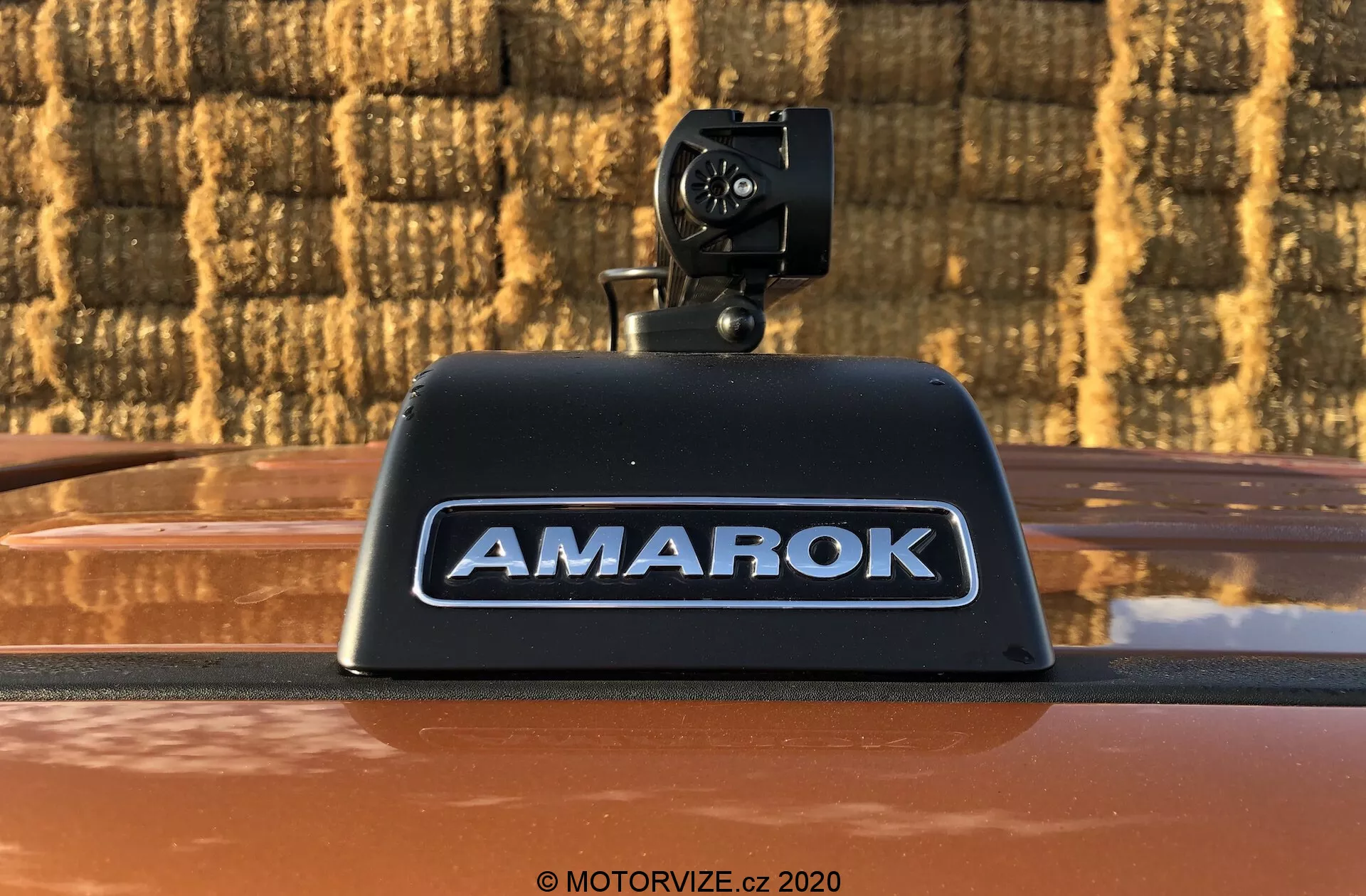 Detailní fotografie pickupu Volkswagen Amarok 2016 zobrazující doplňkovou zpětnou kameru namontovanou na vysokomontážní brzdové světlo s nápisem 'AMAROK' ve vyražených stříbrných písmenech na kovově bronzové karoserii, venku proti pozadí s cihlovým vzorem.