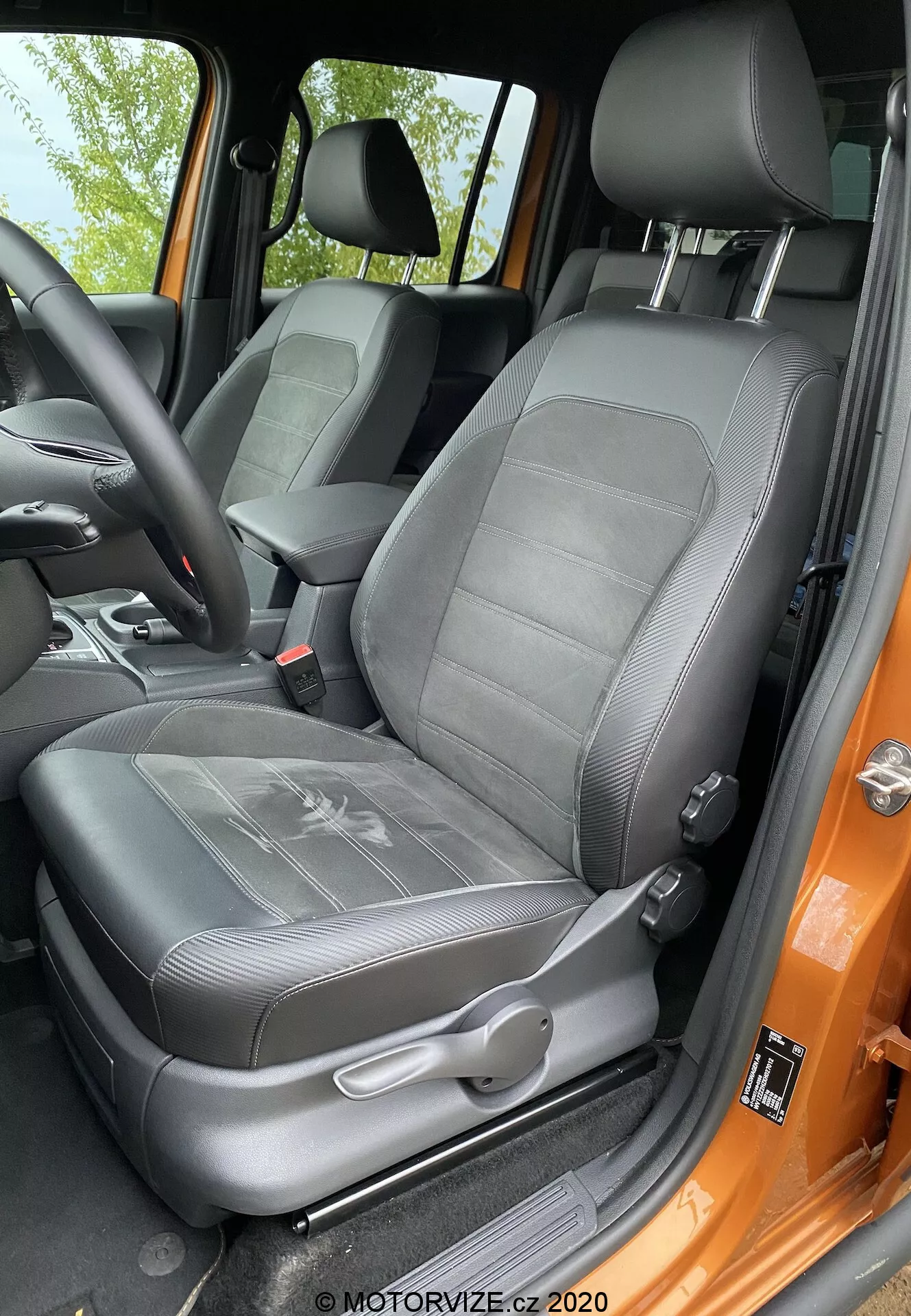 Detailní pohled na sedadlo řidiče modelu VW-nutzfahrzeuge Amarok 2016 z pohledu osoby stojící venku u otevřených dveří. Černé čalouněné sedadlo má vzorovaný design s žebrovým texturováním a integrovanou nastavitelnou opěrku hlavy. Viditelné jsou manuální ovládací páčky a mechanismus tříbodového bezpečnostního pásu. Boční opěrka naznačuje boční podporu a dveřní rám natřený výraznou oranžovou barvou kontrastuje s černým interiérem. Sedadlo vypadá dobře udržované a je osvětlené přirozeným denním světlem.