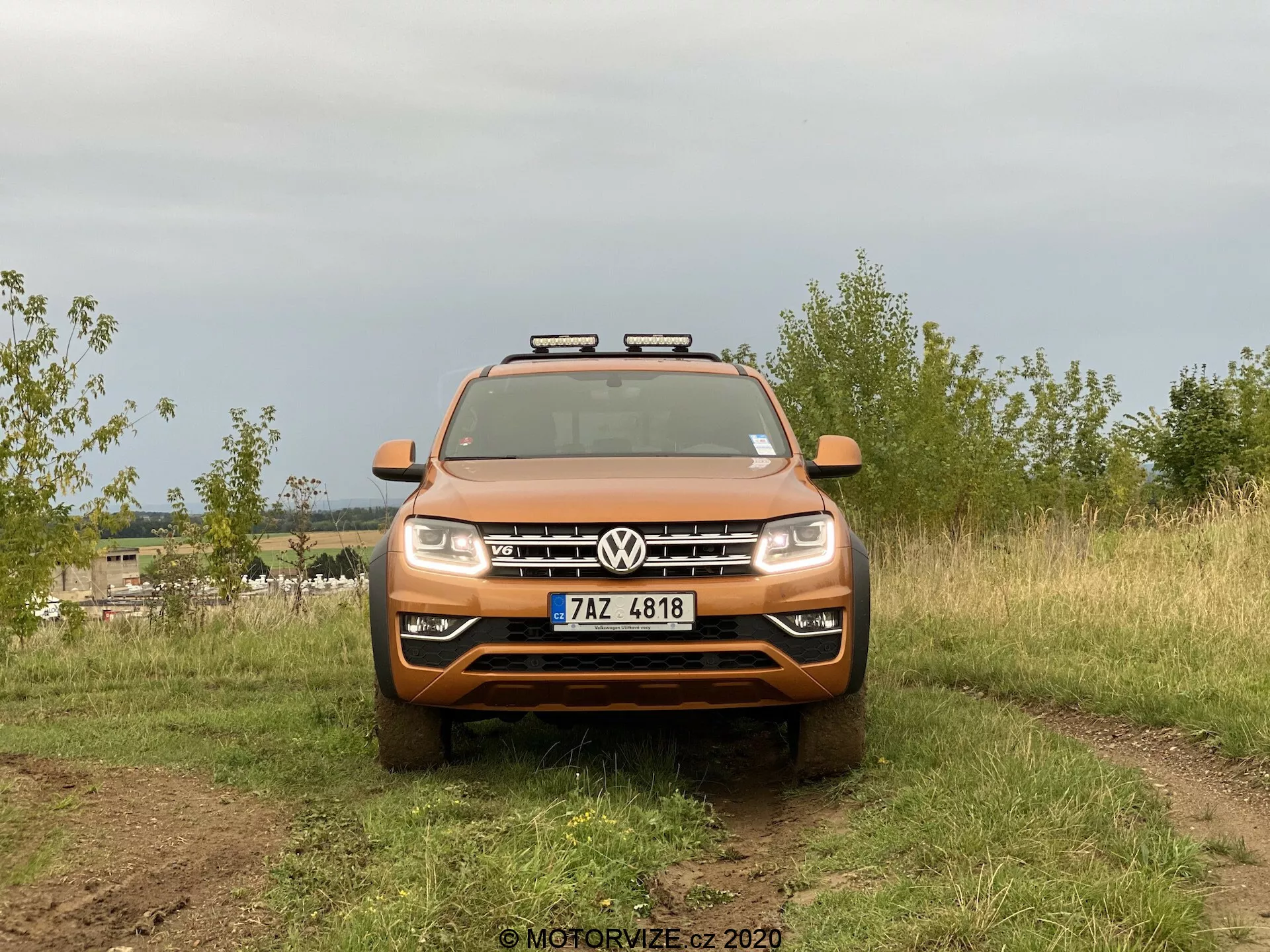 Přední pohled na SUV Volkswagen Amarok zachycený ve venkovním prostředí. Oranžové vozidlo vyniká výrazným logem Volkswagen na masce, horizontálními lamelami, standardními světlomety a přídavnými světly umístěnými na střeše. Obrázek zdůrazňuje terénní schopnosti s částečně viditelnými těžkými pneumatikami a nečistotami na povrchu vozidla. Auto je umístěno v přírodním terénu s rozmazaným pozadím a rozptýleným světlem z oblačné oblohy.