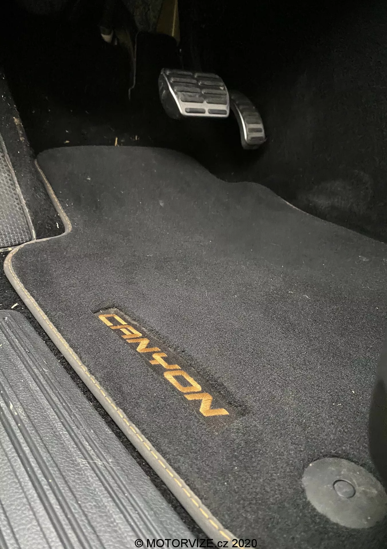 Detailní záběr podlahové plochy na straně řidiče u VW-Nutzfahrzeuge Amarok 2016, zachycující tmavou podložku s vyšitým nápisem 'ENYON'. Viditelné jsou kovové pedály plynu a brzdy, přičemž brzdový pedál je výše, což naznačuje, že je zabrzděno. Podložka je tvarovaná a zajištěná háčkem na pravé straně. Podložka kontrastuje s temnějším okolím, což naznačuje tmavý design interiéru. Scéna je slabě osvětlena, pravděpodobně pořízená v noci nebo za snížených světelných podmínek.