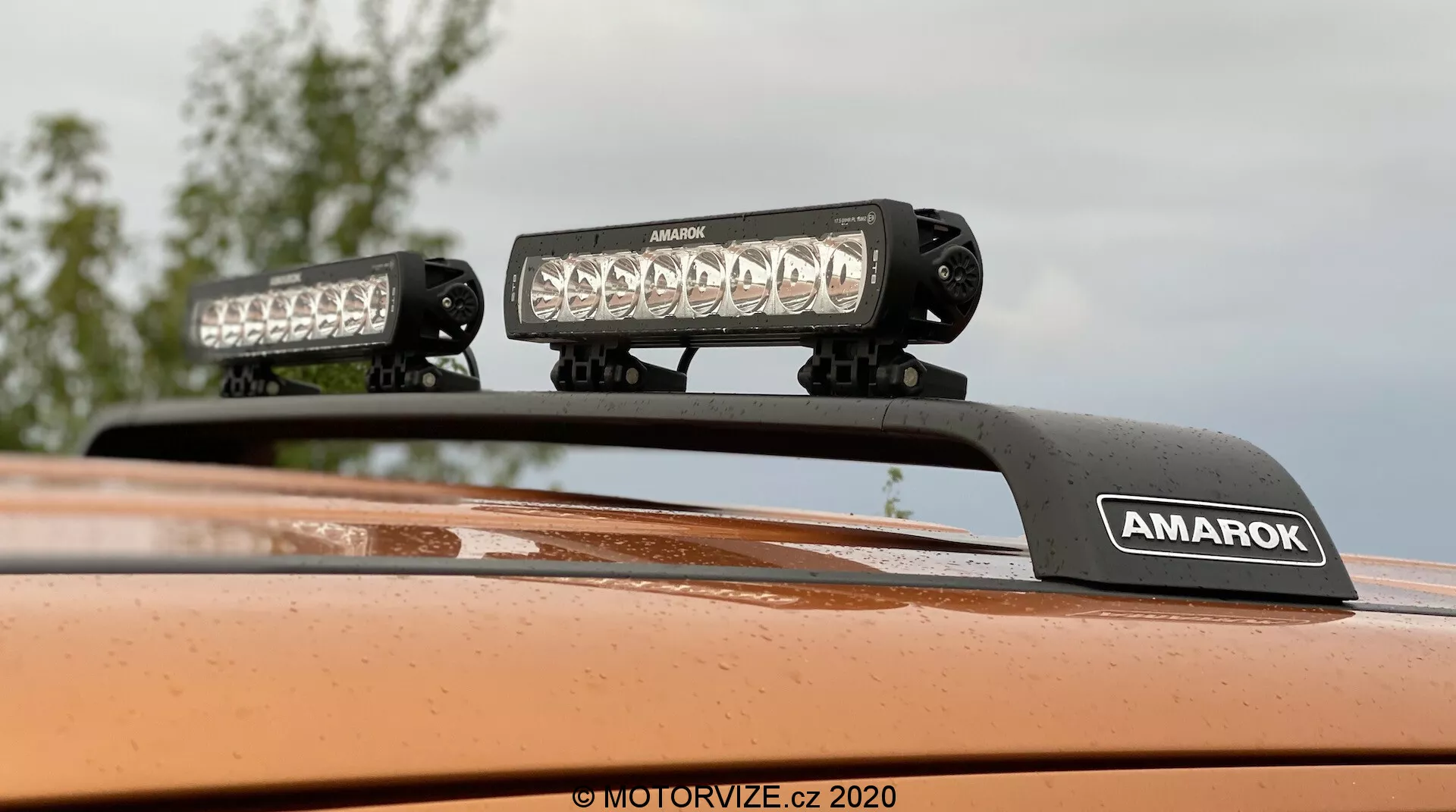 Detailní záběr zespodu na střechu Volkswagenu Amarok 2016 s nástavbovým střechovým osvětlením, dvěma LED světelnými lištami na držáku s nápisem 'AMAROK', metalická oranžová barva; ideální pro doplňky pro terénní vozy nebo osvětlení vozidel.