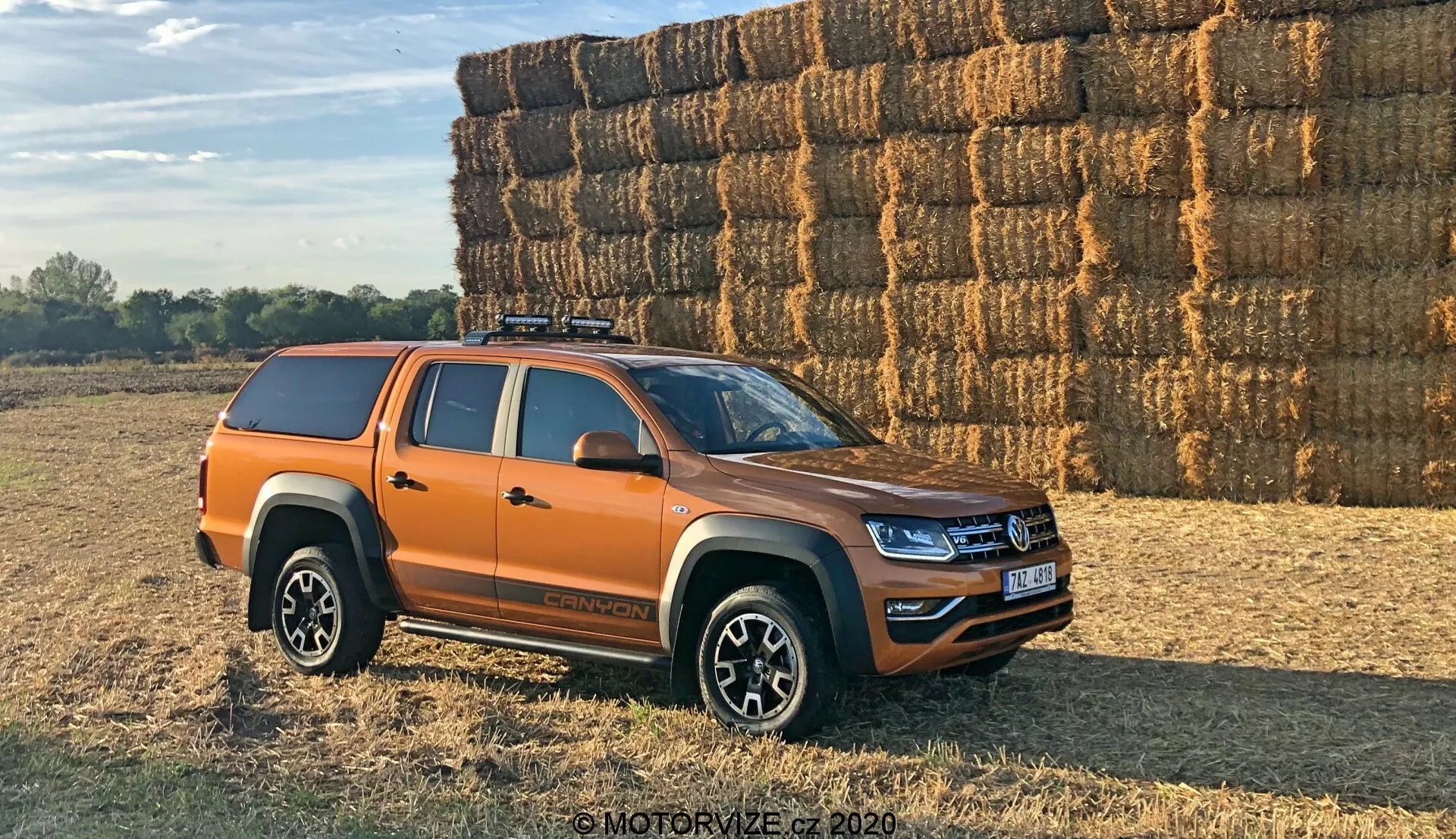 Boční pohled na oranžový pickup Volkswagen Amarok zaparkovaný venku na nerovném, potenciálně hliněném nebo travnatém povrchu. Vozidlo je umístěno pod úhlem 30 stupňů k fotoaparátu, což umožňuje vidět boční profil a částečný pohled na přední masku a světlomety. Moderní pickup střední velikosti má čerstvé designové linie a LED osvětlení, což naznačuje, že se jedná o nedávný model. Viditelné jsou čtyři dveře označující dvojkabinovou konfiguraci, vícepaprsková litá kola, terénní pneumatiky, tvrdý tonnau kryt na nákladovém prostoru a střešní nosič. Boční zrcátka pravděpodobně obsahují ukazatele. Fotografováno ve vřelém denním světle s dlouhými stíny, v pozadí je vidět velká hromada balíků sena, což naznačuje venkovské prostředí.