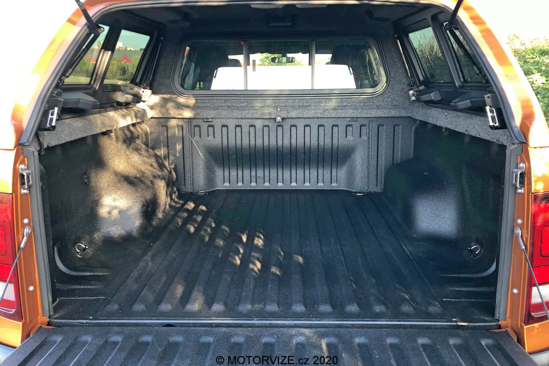 Interiér nákladového prostoru VW Nutzfahrzeuge Amarok 2016, pohled zezadu s otevřenými dveřmi kufru, ukazující zavazadlový prostor. Nákladový prostor obsahuje odolnou vzorovanou výstelku, boční stěny dokončené odolným materiálem, upevňovací body pro zajištění věcí, vnitřní osvětlení pro viditelnost, viditelné zadní sedadla v přední části prostoru a ochranný lem na okraji víka kufru.