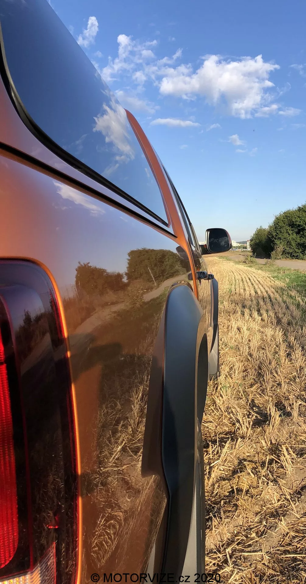 Pohled zblízka na pravou zadní čtvrtku 2016 VW Nutzfahrzeuge Amarok, zvýrazňující lesklou, reflexní kovovou karoserii a integrované zadní světlo s čirou čočkou. Foto, pořízené z nízkého úhlu, ukazuje okenní hranu, černý plastový lem a venkovní sklizené pole s jasnou modrou oblohou.