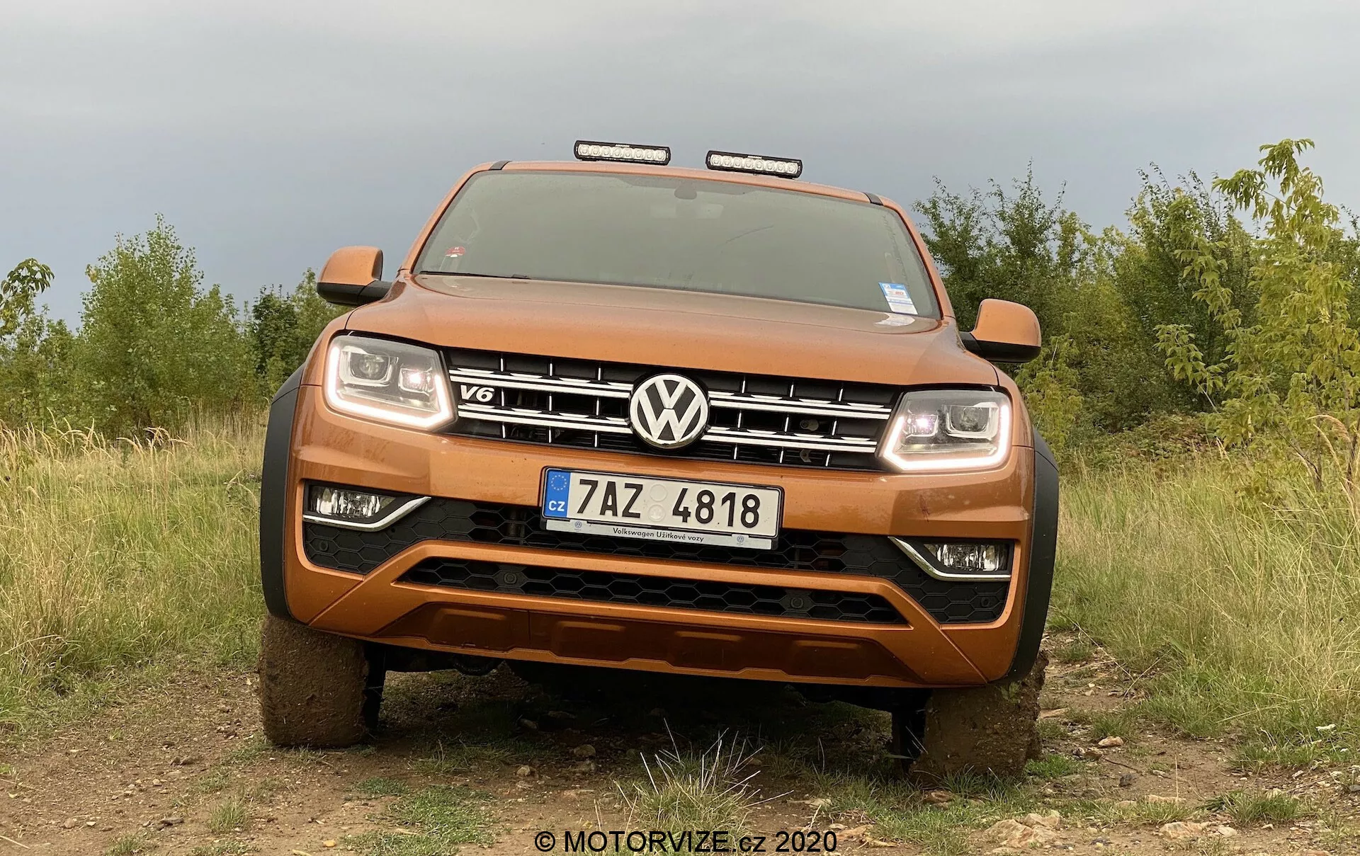 Přední pohled na metalickou oranžovou dodávku Volkswagen Amarok, který ukazuje svou robustnost a terénní schopnosti s výraznou mřížkou s emblémem VW a odznakem V6. Obrázek, pořízený z nízkého úhlu, zachycuje celou šířku přední části vozidla, včetně světlometů a přídavných světel pro terénní jízdu, s přírodním pozadím pole a stromů, které zvýrazňují robustnost pickupu. Viditelné funkce zahrnují pevný přední nárazník, kluznou desku, aktivovaný pokročilý osvětlovací systém a registrační značku České republiky.
