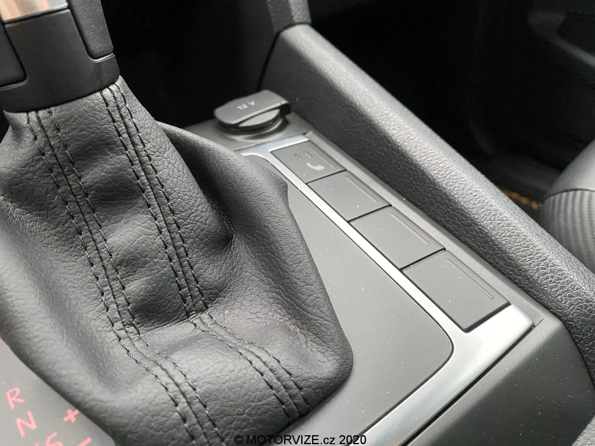 Detailní záběr na středovou konzolu vozu VW Nutzfahrzeuge Amarok 2016, zaměřující se na oblast řazení. Hlavním prvkem je řadicí páka, potažená černou kůží s viditelným šitím. Písmena 'PRNDS' za řadicí pákou označují automatickou převodovku. Napravo jsou čtyři čtvercová tlačítka zarovnaná s povrchem konzoly, pravděpodobně ovládající různé funkce vozu. Oblast za tlačítky může sloužit jako malý úložný prostor. Materiály a kvalita provedení naznačují vysokou úroveň řemeslného zpracování. Ambientní světlo z levého horního rohu zvýrazňuje detaily.