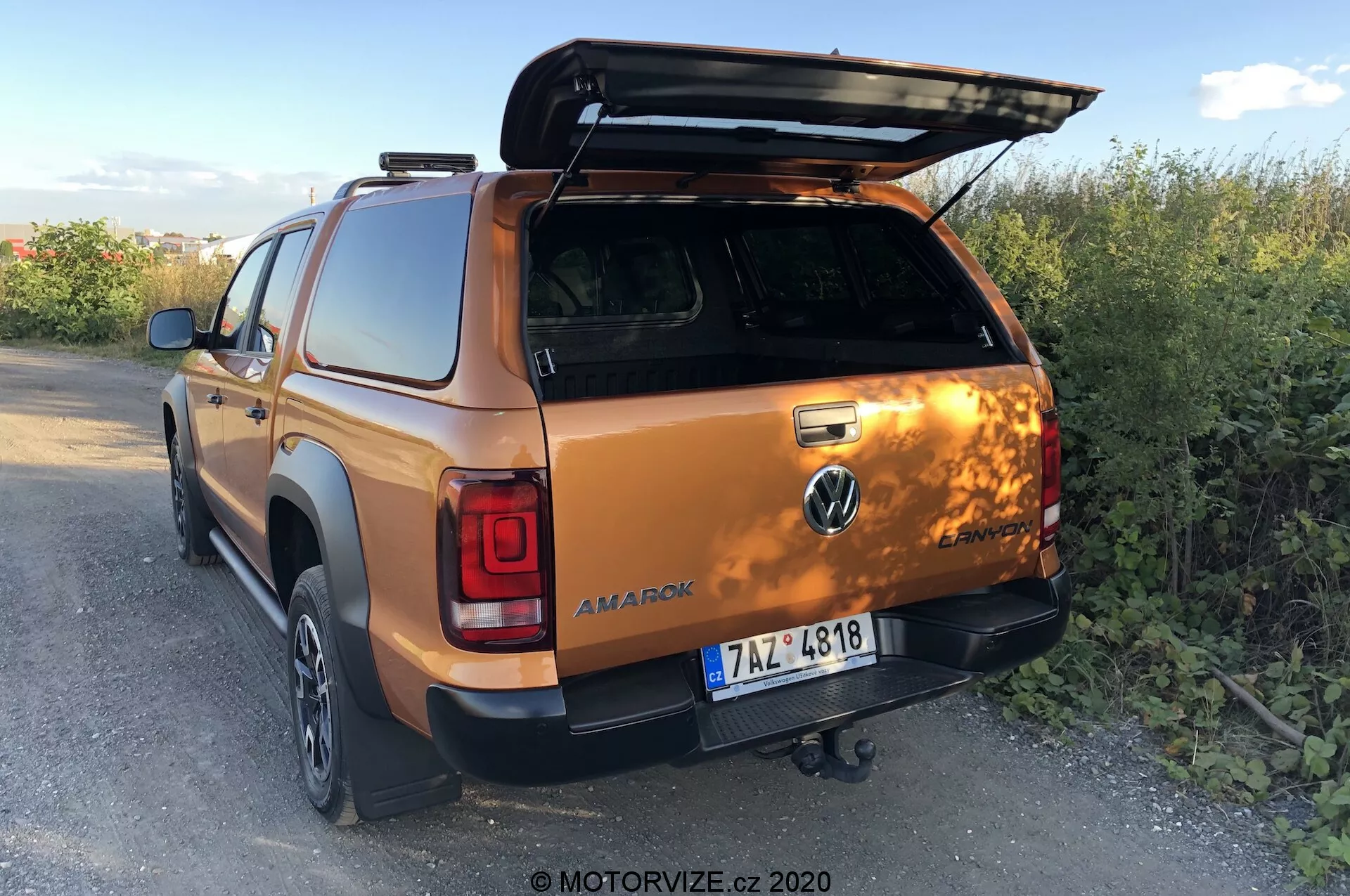 Pohled na Volkswagen Amarok 2016 zezadu a ze tří čtvrtin, ukazující otevřený kryt zavazadlového prostoru, tažné zařízení, dveře druhé řady kabiny a část pravého zadního kola a nárazníku, zasazený do venkovního venkovského prostředí za soumraku. Foto od MOTORVIZE.cz, 2020.