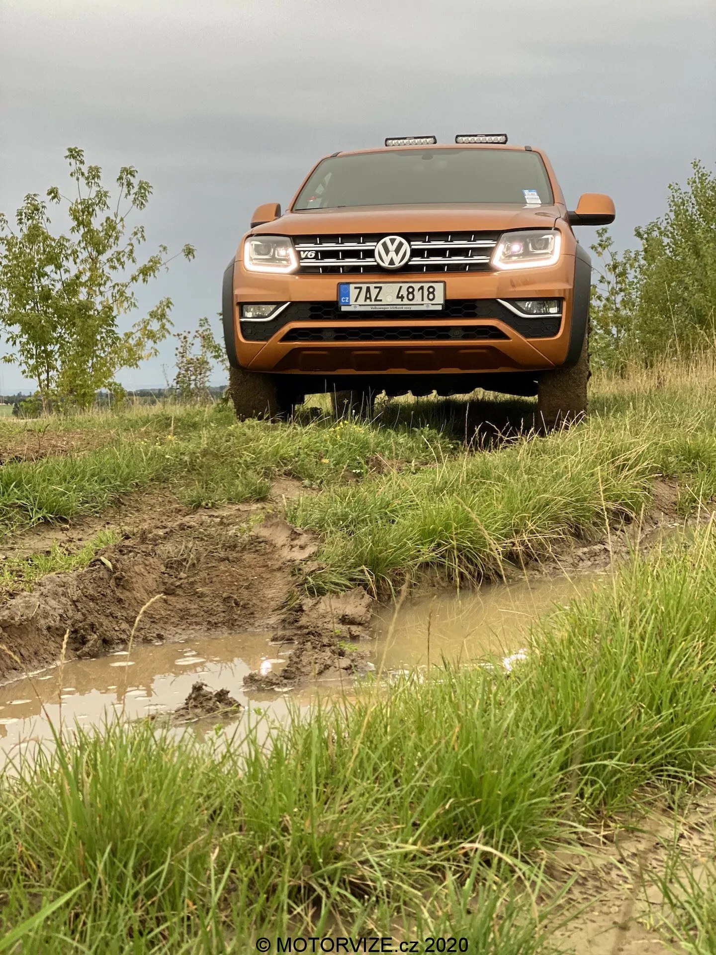 Přední pohled na oranžové terénní vozidlo Volkswagen Amarok 2016, zdůrazňující přední mřížku, nárazník, kapotu a sestavu světlometů. Zachyceno z nízkého úhlu, aby se zdůraznil zvýšený postoj vozu, obraz ukazuje drsnou, blátivou cestu s vodními loužemi a špinavou exteriér, což zdůrazňuje jeho terénních schopnosti. Vozidlo disponuje výraznou mřížkou Volkswagen s emblémem, obdélníkovými světlomety a pomocným osvětlením nad mřížkou. SPZ je viditelná na robustním předním nárazníku, zatímco dlouhá plochá kapota podtrhuje velikost a sílu Amaroku.