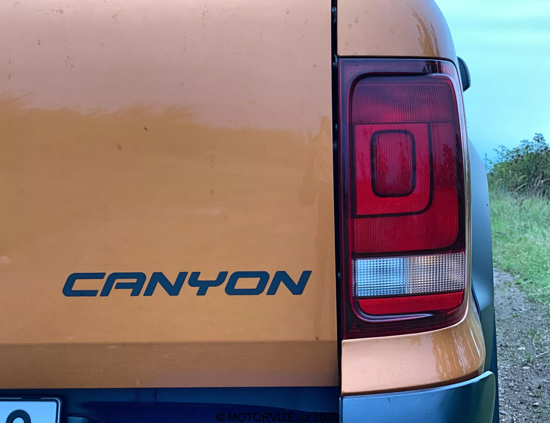 Detailní pohled na pravý zadní roh vozu VW Nutzfahrzeuge Amarok 2016. Na zadních dveřích je vidět odznak 'CANYON' uprostřed známek lehkého znečištění, což naznačuje použití na nezpevněných cestách. Fotografie zdůrazňuje především moderní obdélníkový světelný klastr se sekcemi pro brzdová, zpětné a pravděpodobně směrová světla. Snímek pořízený přibližně pod 45stupňovým bočním úhlem zahrnuje částečně viditelnou karoserii vozu s venkovským nebo polovenkovským pozadím.