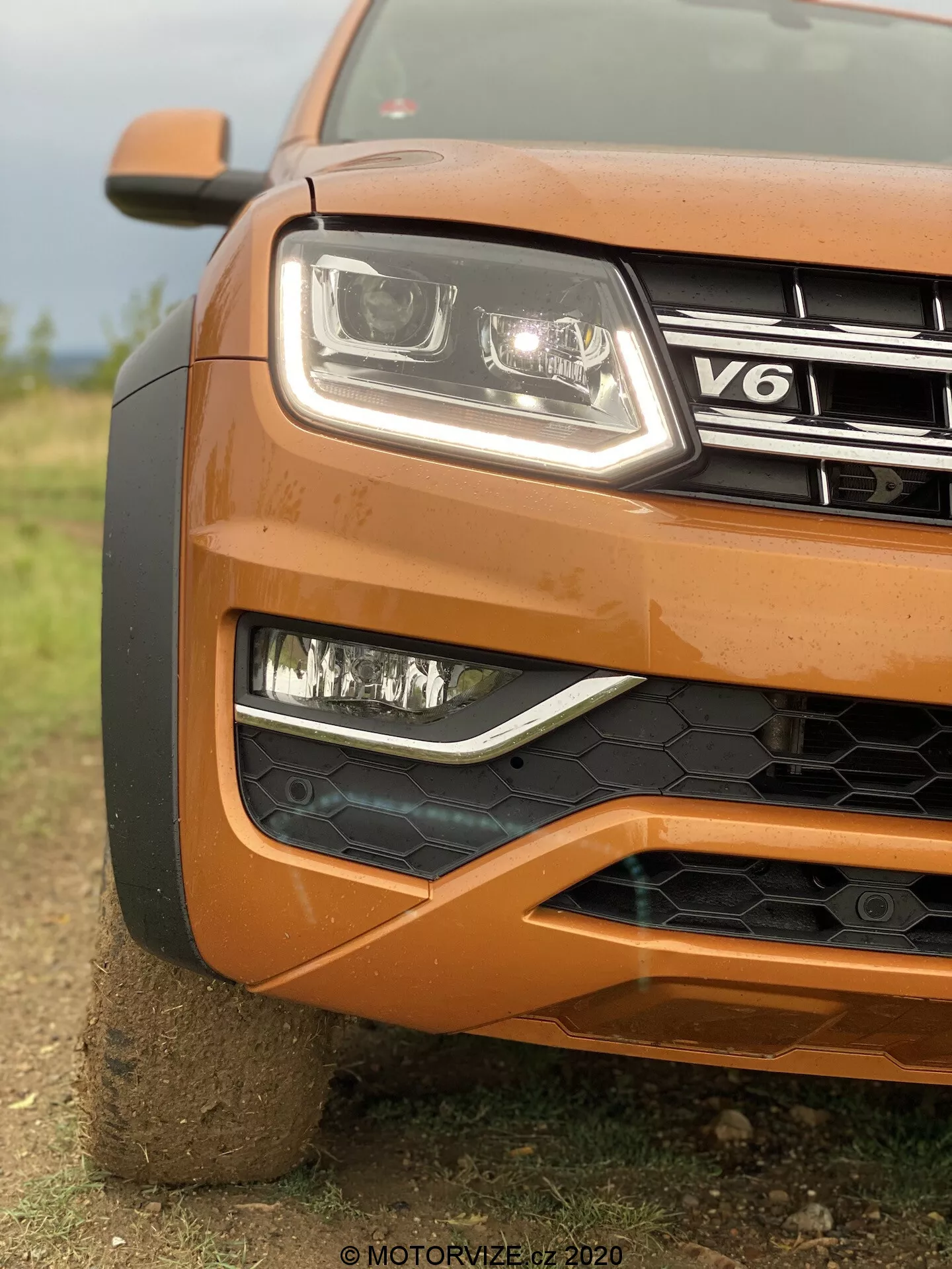 Detailní záběr na levý přední roh oranžového VW Nutzfahrzeuge Amarok 2016, zobrazující světlomet s moderním čirým krytem a LED technologií, mlhovky v černé mřížce, částečně viditelnou mřížku se znakem V6, nárazník s černou voštinovou strukturou, tvarovanou kapotou a částečně viditelným velkým kolem, s rozmazaným pozadím zdůrazňujícím texturu a robustní design vozu