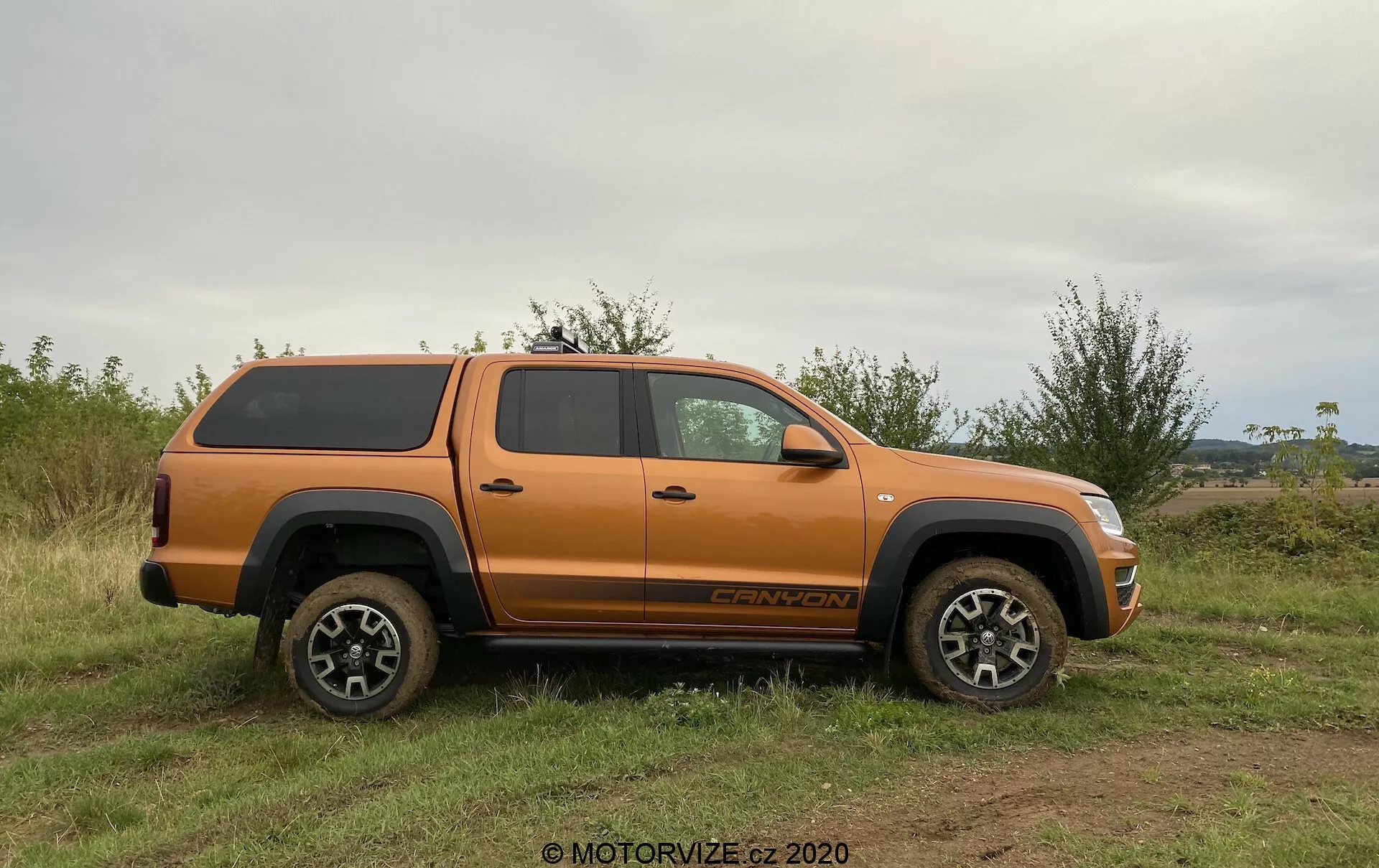 Boční profil oranžového pickupu VW Nutzfahrzeuge Amarok 2016 s tvrdou krycí plachtou, zaparkovaného na nerovném travnatém terénu. Vozidlo má vysokou světlou výšku, odolná kola a pneumatiky s vícepaprskovým designem z lehkých slitin, vhodné pro terénní použití. Viditelné prvky zahrnují boční zrcátka, kliky dveří, blatníky, stupačku, prostornou kabinu se čtyřmi dveřmi, odznak na předním kole a název výbavy nad zadním kolem. Přirozené světlo a vyvážený úhel zvýrazňují detaily vozidla bez ostrých stínů, zdůrazňují jeho terénní schopnosti a bezpečné uložení nákladu.
