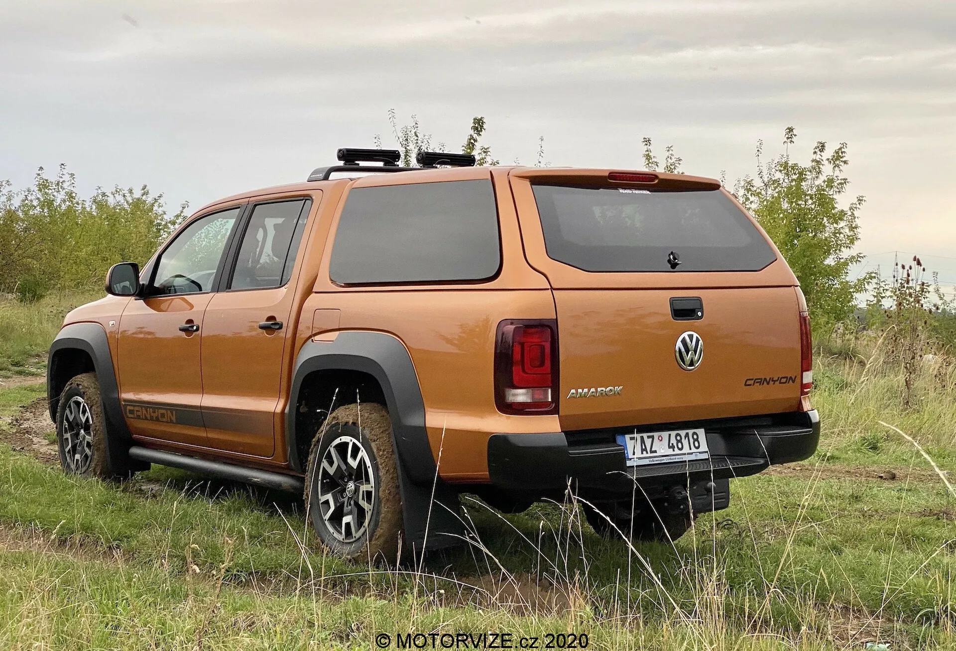 Tříčtvrteční pohled zezadu na oranžový pickup Volkswagen Amarok 2016, zobrazující design kol, boční profil, zadní okno, zadní světla, robustní zadní nárazník a pevný kryt nákladového prostoru, zaparkovaný na nezpevněném povrchu s vegetací, zdůrazňující jeho terénní schopnosti.