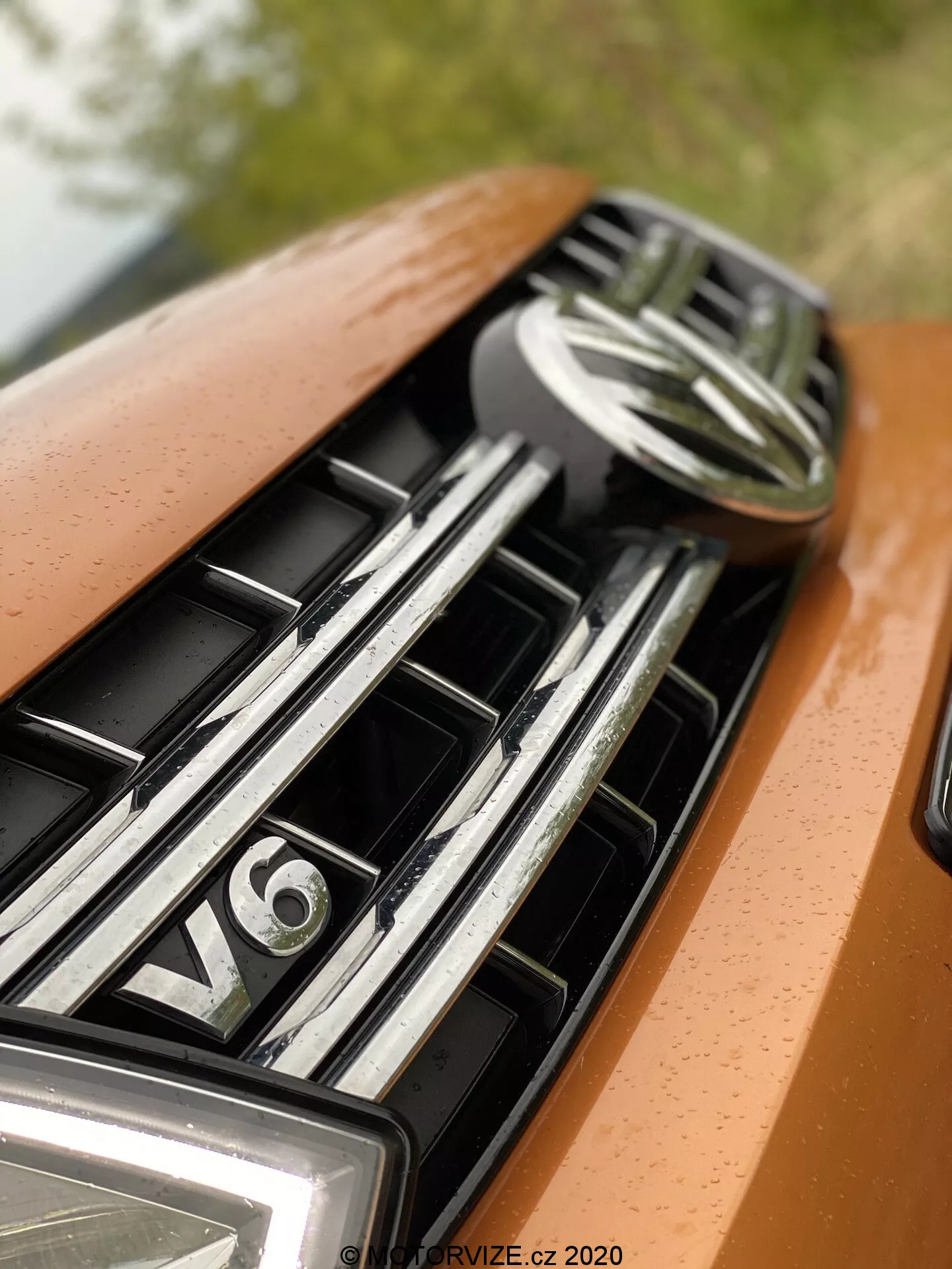 Detailní záběr na kapotu a masku auta, zaměřený na horní část s centrálním znakem a odznakem motoru V6. Maska obsahuje vodorovné, lesklé chromové lamely, což naznačuje luxusní vozidlo. Lak auta má metalickou oranžovou nebo bronzovou barvu a měkké, rozptýlené osvětlení zdůrazňující hladký design a prémii.