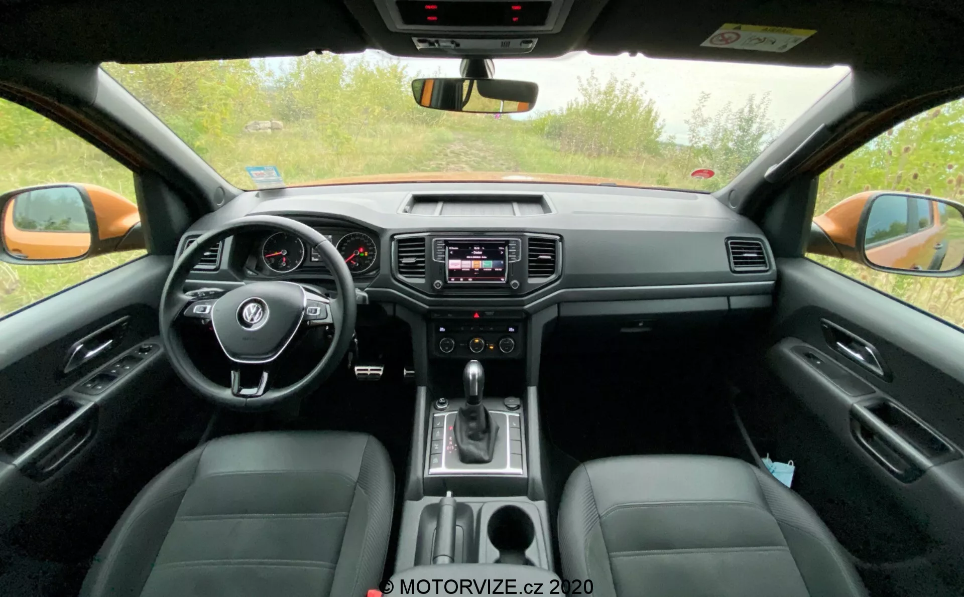 Přední část interiéru vozu 2016 VW-Nutzfahrzeuge Amarok. Snímek pořízený z perspektivy zadního sedadla ukazuje středové řízení s ovladači, analogový přístrojový panel s malým digitálním displejem, infotainment systém na středové palubní desce s ovládáním audia a klimatizace, řadicí páku indikující automatickou převodovku a kožená sedadla. Obrázek také zachycuje zpětné zrcátko, vnější zpětná zrcátka a přední postranní okna, čímž zdůrazňuje jak technologické, tak komfortní prvky.