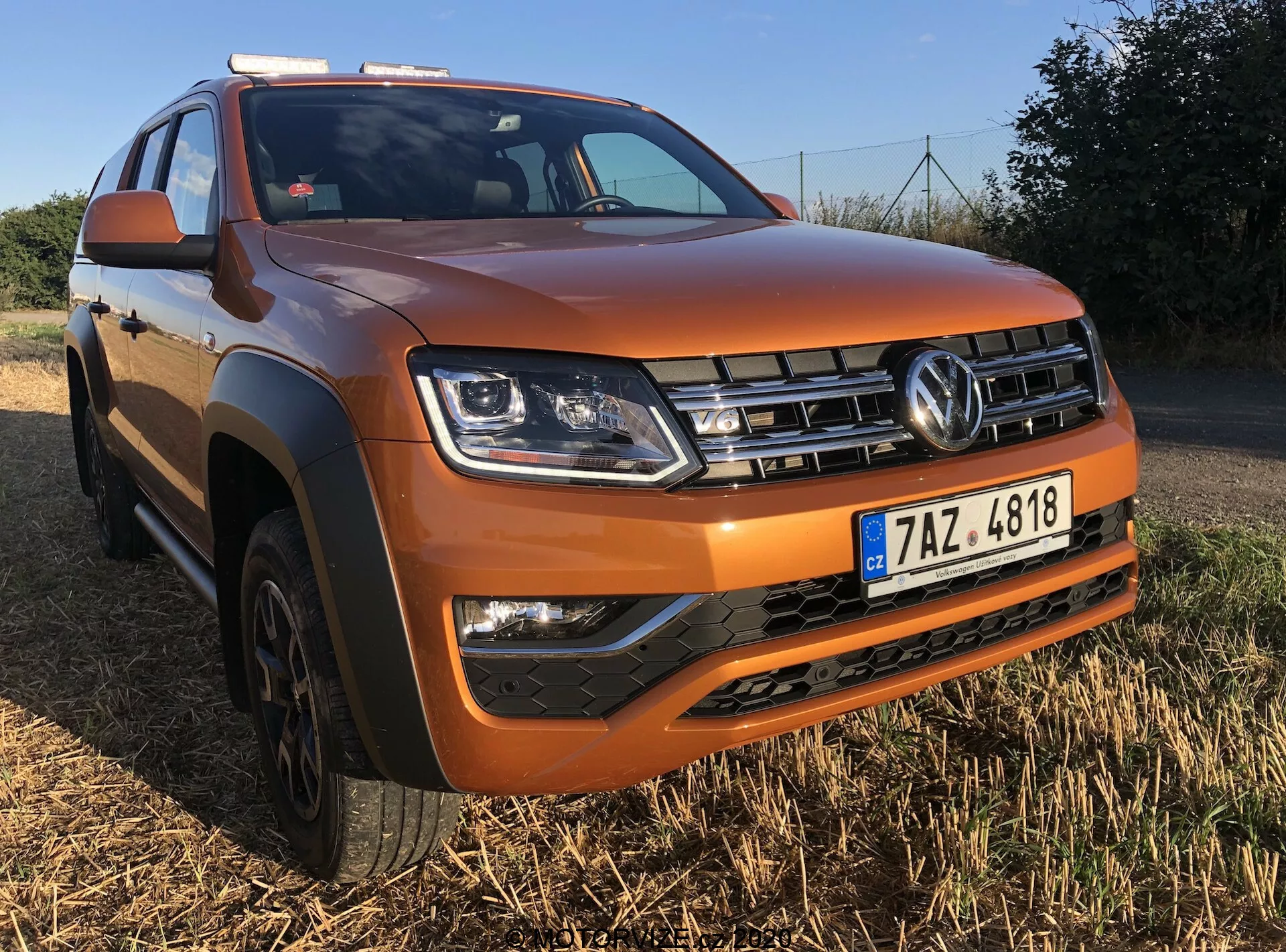 Obrázek Volkswagen Amarok 2016 zaparkovaného na travnaté ploše s jasnou oblohou v pozadí. Snímek z nízkého úhlu zepředu šikmo na stranu řidiče zvýrazňuje přední mřížku s logem Volkswagen, hranaté světlomety, mlhovky v černé spodní části nárazníku a vyvýšenou středovou kapotu. SUV je lakováno v metalické oranžové a má černé plastové lemování na obloukech kol a ochranný kryt na předním nárazníku. Viditelné jsou stříbrné střešní ližiny a pětiramenné alu kola, což zdůrazňuje jeho robustní a do terénu připravený design.