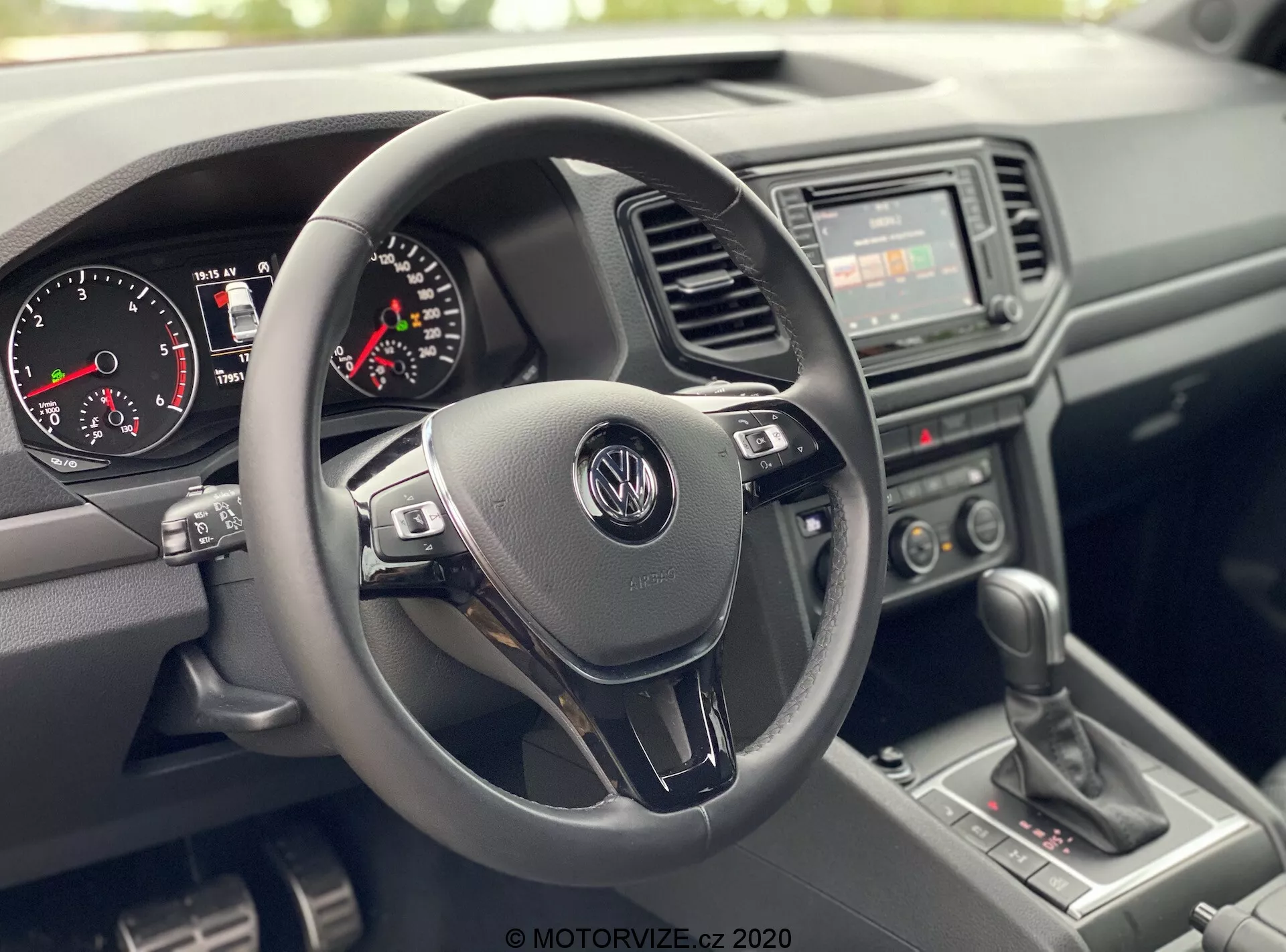 Pohled do interiéru VW Nutzfahrzeuge Amarok 2016 z perspektivy předního spolujezdce, zobrazující řidičskou kabinu. Ústřední pozornost na tříramenný volant s logem Volkswagen a multifunkčními ovládacími prvky, přístrojový panel s analogovým tachometrem, otáčkoměrem a malým digitálním displejem, základní rádio s manuálními ovládacími prvky klimatizace na středové konzole, manuální řadicí páku a texturované plastové materiály pro přístrojovou desku a dveře, zdůrazňující odolný a praktický design.