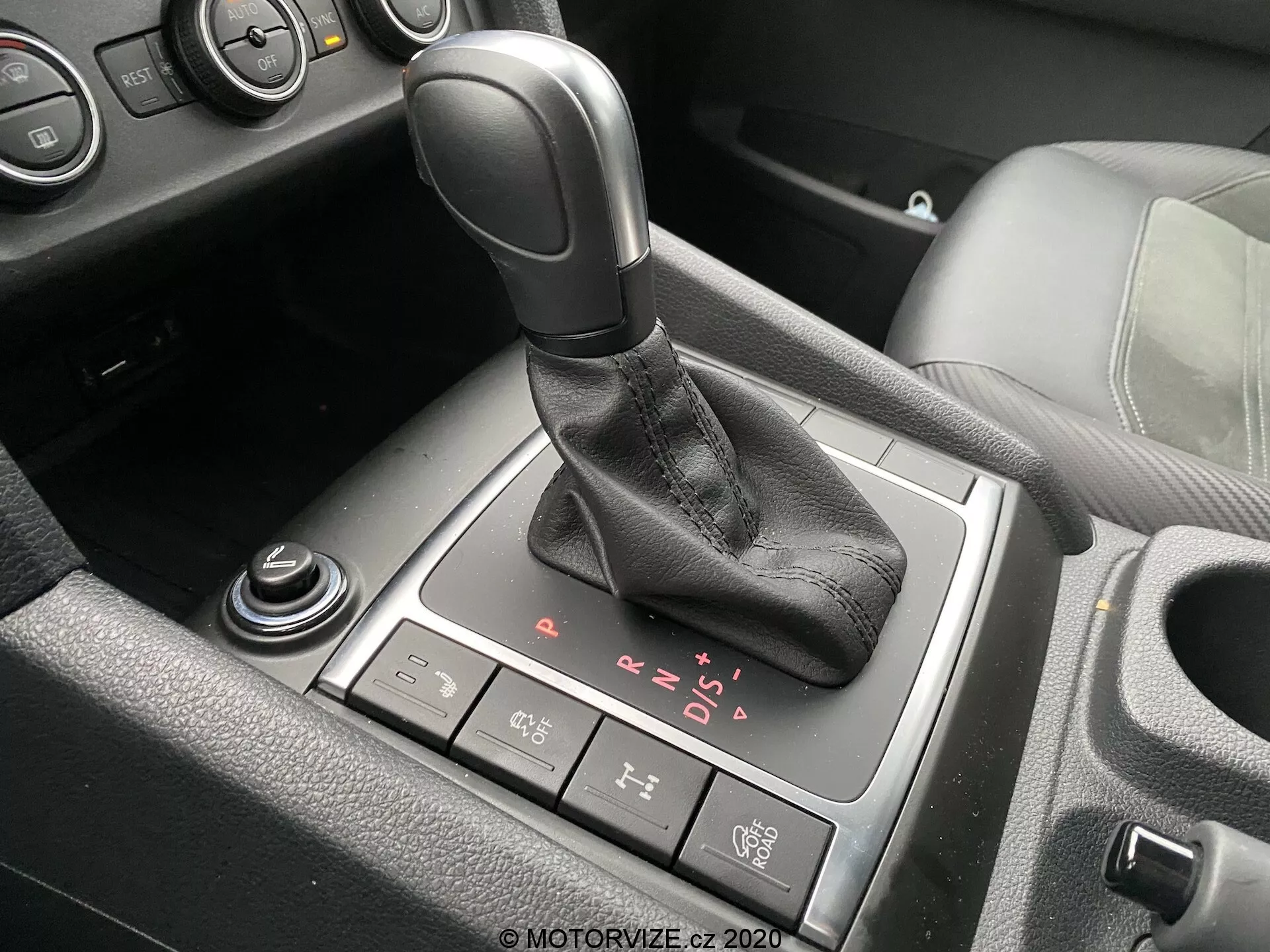 Detailní pohled na oblast řazení v interiéru VW Nutzfahrzeuge Amarok 2016. Automatická řadicí páka s černou rukojetí a koženým návlekem je umístěna uprostřed obrazu s viditelnými možnostmi P, R, N, D a sekvenčního manuálního režimu. Tlačítko elektronické ruční brzdy je umístěno za řadicí pákou, zatímco částečně viditelný rotační volič a další volič se nachází vpředu a vlevo dole. Částečné pohledy na držák na poháry a středovou konzoli s černým plastem a hliníkovými nebo chromovanými akcenty také naznačují moderní vozidlo se střední nebo vyšší úrovní výbavy.