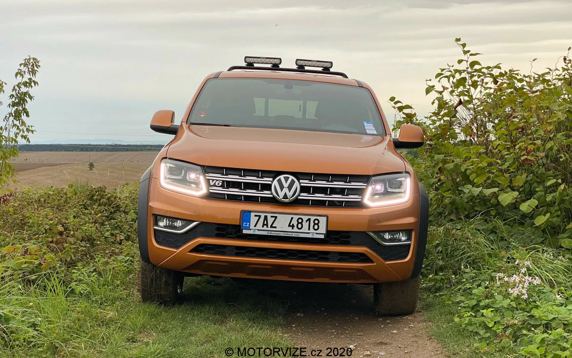 Pohled zpředu na Volkswagen Amarok SUV v metalické oranžové barvě, zaparkovaný na trávě s venkovským off-road pozadím, ukazující symetrickou masku, světlomety, chromové akcenty a pomocná střešní světla.