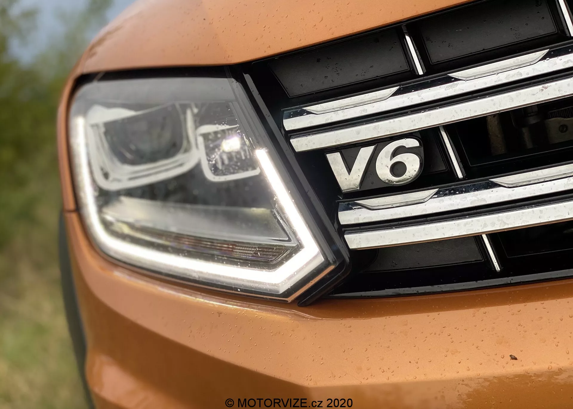 Blízký záběr na přední levou část vozidla VW Nutzfahrzeuge Amarok 2016. Obrázek zachycuje moderní, úhlový světlomet s LED denním svícením a technologií projektorového paprsku spolu s chromovými detaily. Mřížka má horizontální chromované proužky a štítek „V6“, což znamená, že vůz má šestiválcový typ motoru. Část kapoty se objevuje v živé oranžové barvě, což naznačuje sportovní design.