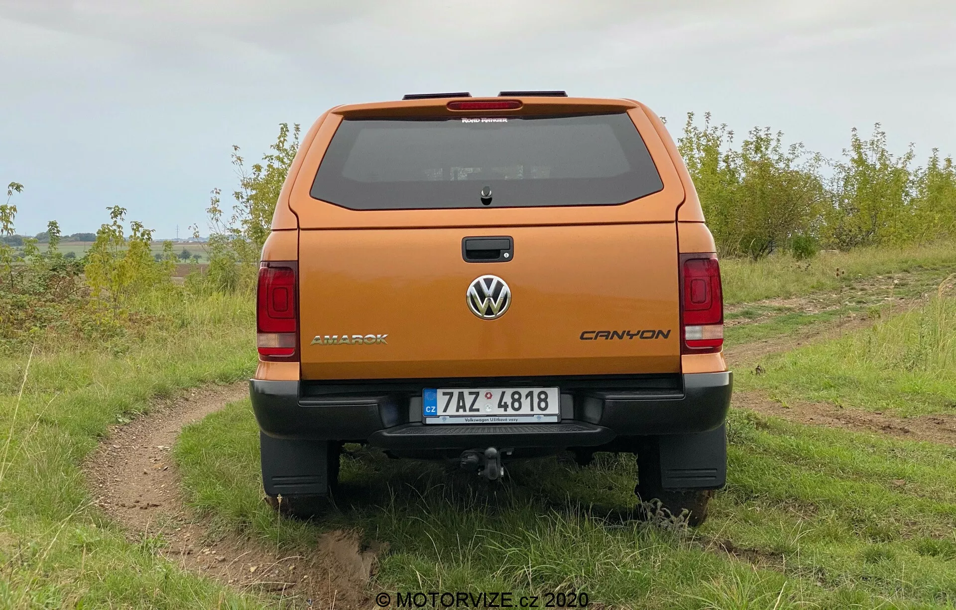 Pohled zezadu na metalickou oranžovou Volkswagen Amarok 2016 pickup zaparkovaný venku na nerovném štěrkovitém cestě s travou po obou stranách, pod zataženou oblohou. Fotografie ukazuje jasnou perspektivu zadní části vozu včetně odznaku Volkswagen, jmenovky Amarok a znaku V6. Vozidlo má zadní nárazník s integrovanou schůdkem, centrálně umístěnou poznávací značkou a viditelným tažným zařízením. Viditelná jsou vertikálně orientovaná koncová světla a třetí brzdové světlo na střešní linii nad zadním oknem, stejně jako odmrazovací linky.