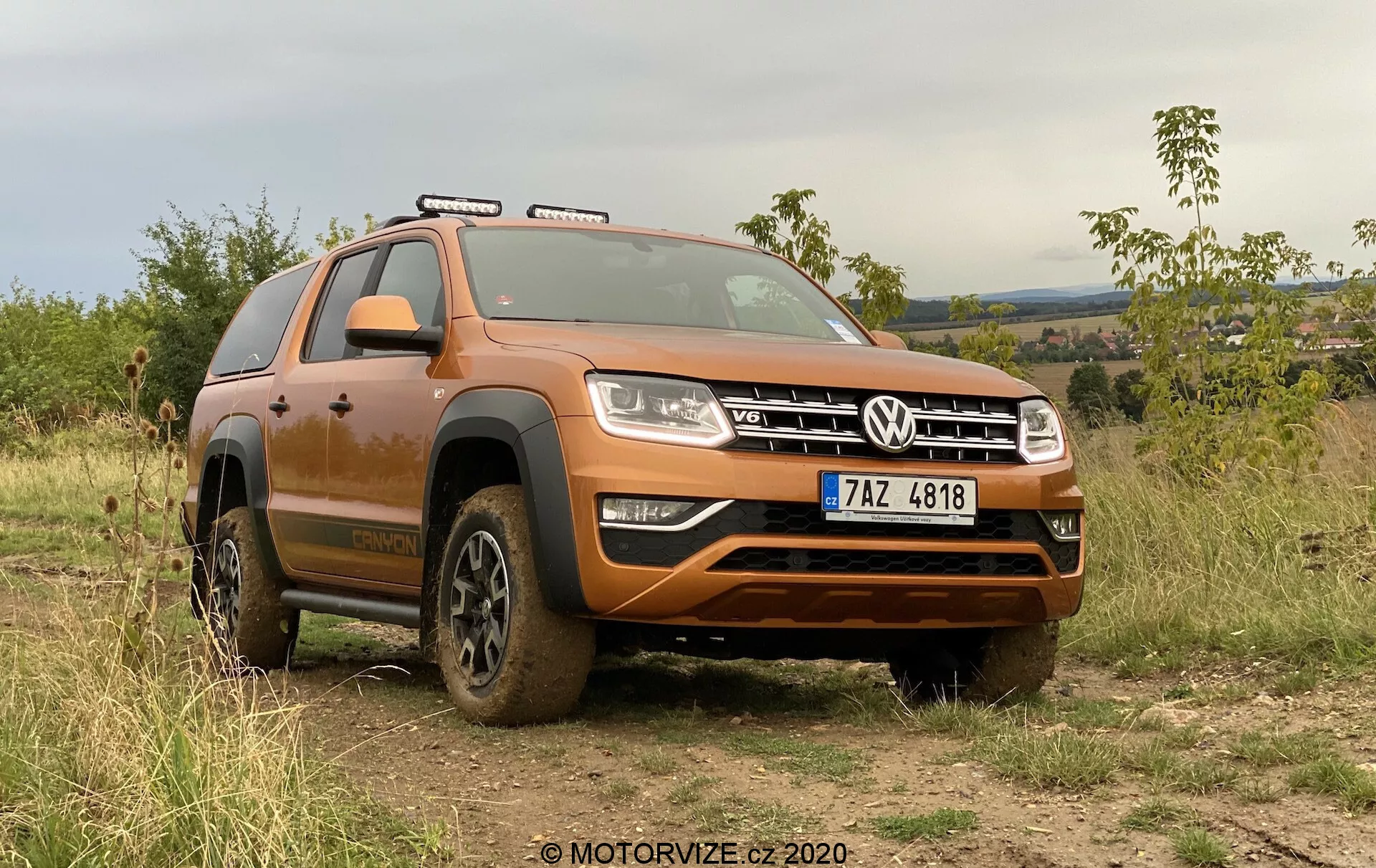 Obrázek zobrazuje moderní SUV, model Volkswagen Amarok 2016, zaparkovaný venku na nerovném terénu, který ukazuje jeho schopnosti pro jízdu v terénu. Pohled z nízkého čelního úhlu odhaluje přední čtvrtinu vozidla pokrytou bahenními zbytky z nedávného terénního využití. Široká přední mřížka s charakteristickým znakem VW je lemována obdélníkovými světlomety s LED denními světly. Pod mřížkou je robustní přední nárazník s ochranou spodní části karoserie. Konturovaná kapota přidává SUV svalnatý vzhled. Střešní ližiny naznačují možnost přepravy dalšího nákladu. Kola částečně zahalená bahnem mají hluboký dezén pro lepší trakci v terénu. Viditelný je výrazný oblouk nad předním kolem a nášlap, který usnadňuje vstup a výstup z vozu. Viditelné jsou také střešní osvětlení a boční zrcátka. Auto je zasazeno do pozadí s vegetací a kopcovitou krajinou pod zamračenou oblohou, což zdůrazňuje jeho terénní schopnosti a venkovní přitažlivost. Levé řízení a viditelná registrační značka naznačují, že se vozidlo nachází v zemi, kde jezdí vpravo. Tento záběr zdůrazňuje design vozu, terénní prvky a moderní osvětlovací technologie.