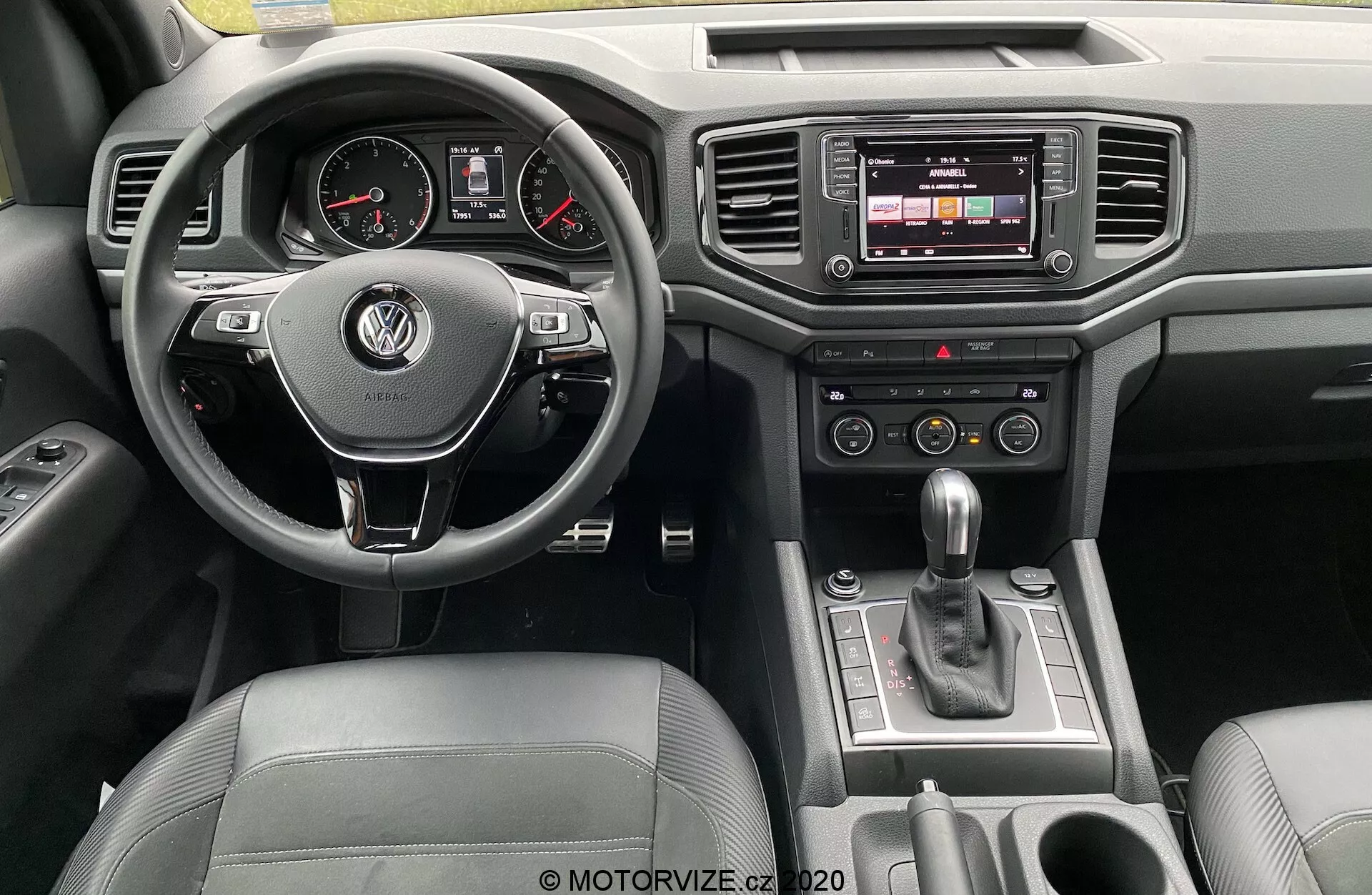 Pohled na přístrojovou desku Volkswagenu Amarok 2016 z pohledu řidiče. Centrálně umístěný multifunkční volant potažený kůží s logem VW, analogovými ciferníky a digitálním displejem v přístrojové desce. Centrální konzole obsahuje dotykový infotainment systém, tlačítka klimatizace a automatickou řadicí páku. Luxusní detaily zahrnují kůži a chromované povrchy, s viditelnými ovladači na dveřích.