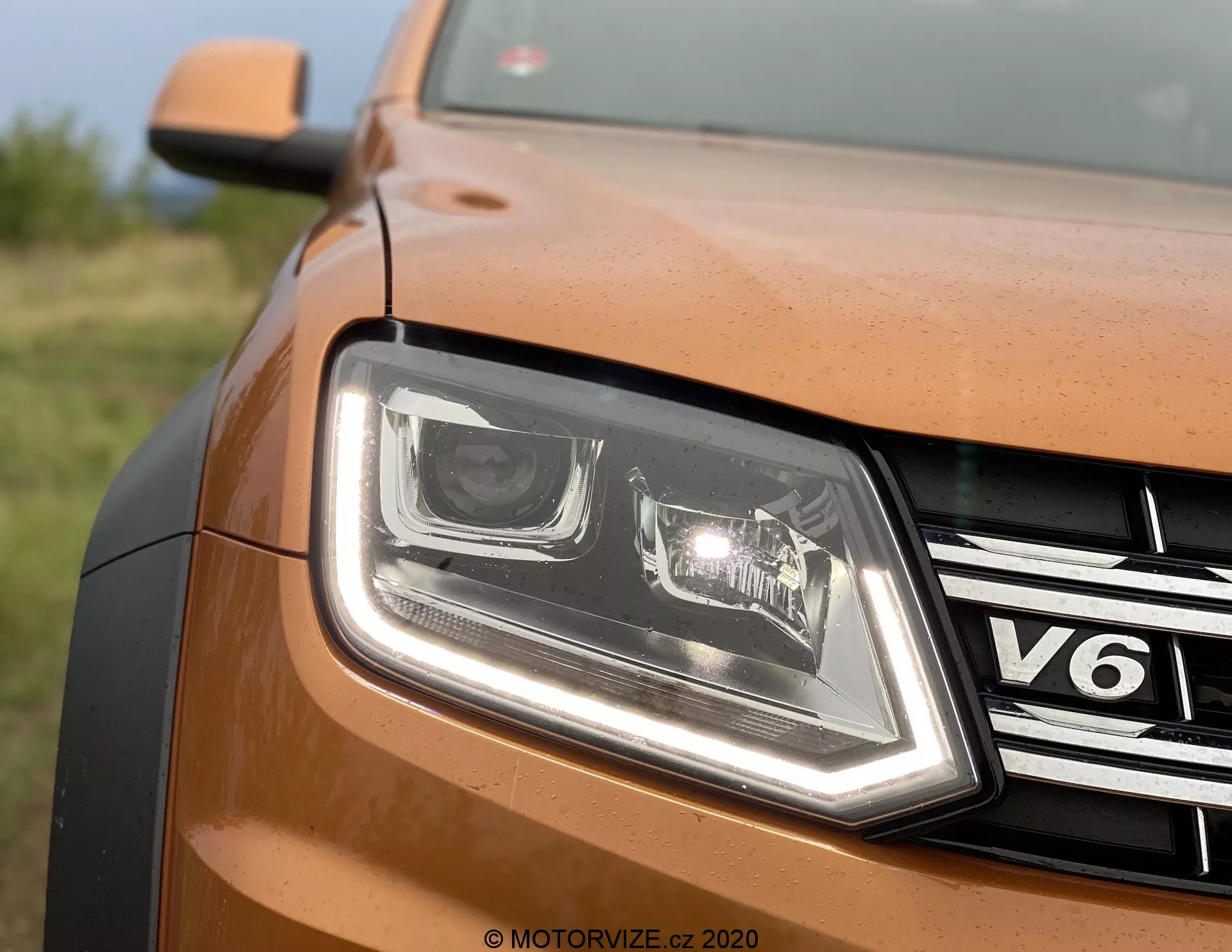 Detailní záběr čelního levého pohledu na bronzově nebo měděně zbarvené auto VW Nutzfahrzeuge Amarok 2016. Zobrazuje LED světlomety s výraznými denními světly, chromovou mřížku s nápisem 'V6', metalický lak karoserie, hranu kapoty a vodoznak 'MOTORVIZE.CZ 2020', což naznačuje původ z českého recenzního webu.