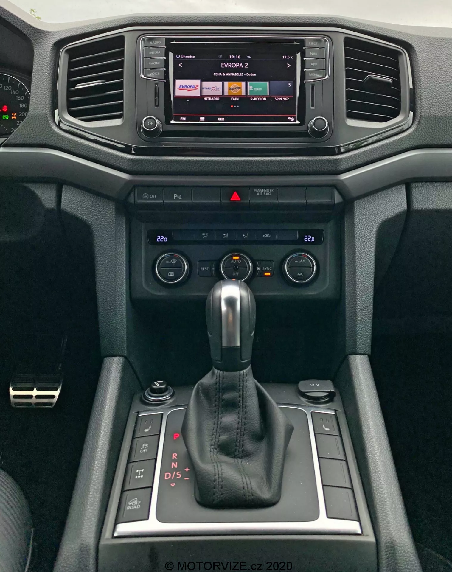 Detailní pohled na moderní středovou konzoli VW Amarok 2016 se zaměřením na infotainment systém a řadicí páku. Infotainment displej naznačuje dotykové ovládání, možná zobrazuje rádio nebo mediální výstup. Níže jsou vodorovné větrací otvory obklopené měkkými materiály s texturovaným designem. Pod větráky jsou ovládací prvky klimatizace a spínače pro nastavení teploty a proudění vzduchu. Dominuje automatická řadicí páka v režimu 'P', což naznačuje automatickou převodovku s možnostmi ručního řazení. Vedle řadicí páky jsou malé úložné přihrádky a otočný ovladač pravděpodobně pro infotainment nebo systém řízení jízdních režimů. Design interiéru zdůrazňuje jak funkčnost, tak pohodlí.