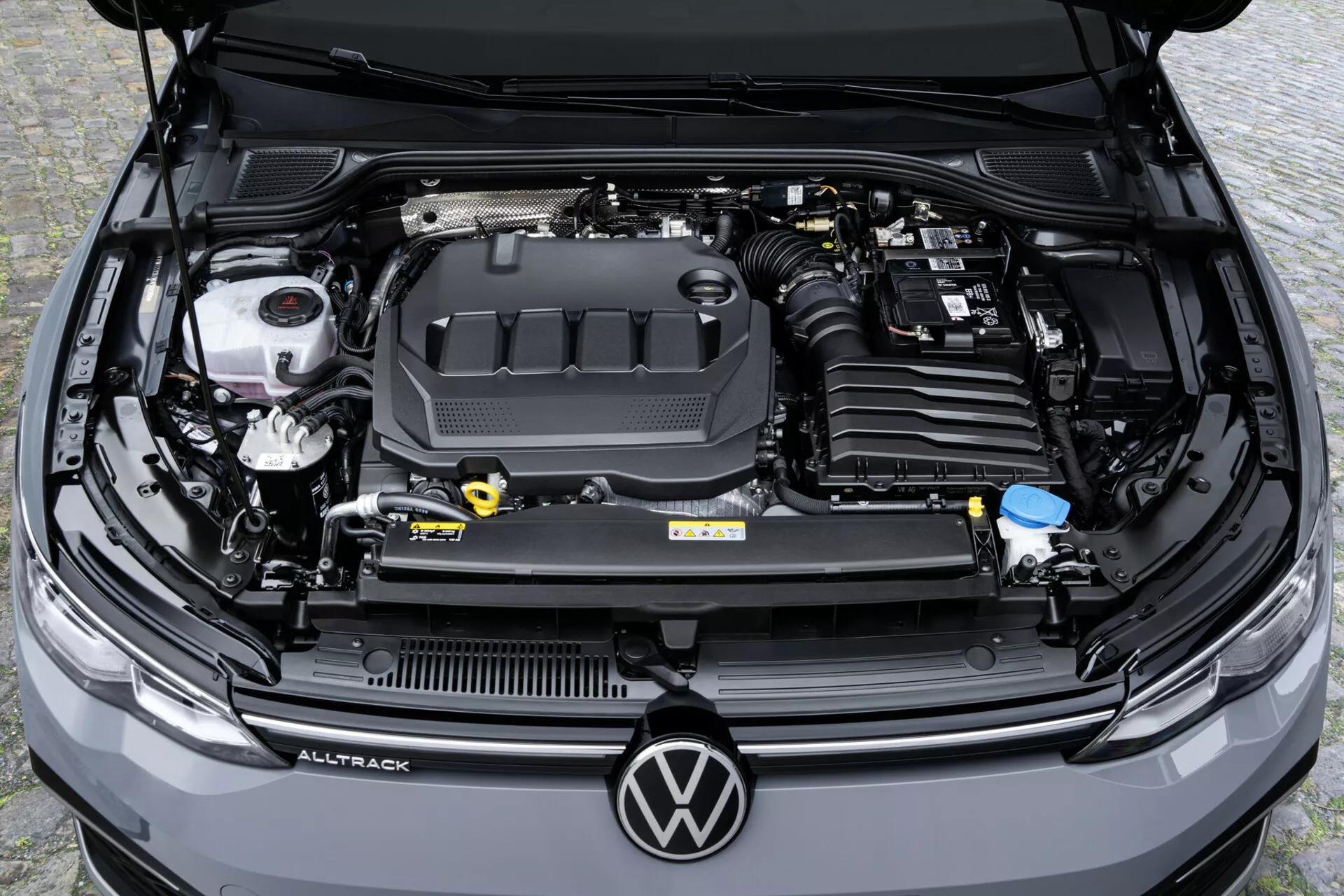 Pohled shora na otevřený motorový prostor VW Golf Alltrack 2017 zdůrazňující mechanické a technologické komponenty včetně krytu motoru s označením značky, nádrže na chladicí kapalinu, loga značky na mřížce chladiče, viditelných hadic, svazků kabelů, nádoby na kapalinu do ostřikovačů, nádrže na brzdovou kapalinu a filtru vzduchu