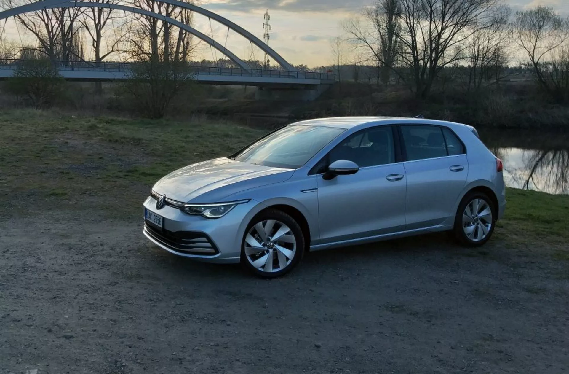 Volkswagen Golf GTD z roku 2017 v barvě stříbrné, zobrazený z frontálního tříčtvrtečního pohledu, který zdůrazňuje jeho elegantní vodorovná LED světlomety, minimalistickou uzavřenou mřížku, charakteristická slitinová kola a aerodynamické linie karoserie, zaparkovaný na nezpevněné ploše s klidnou vodní hladinou a obloukovým mostem na pozadí během zlaté hodiny