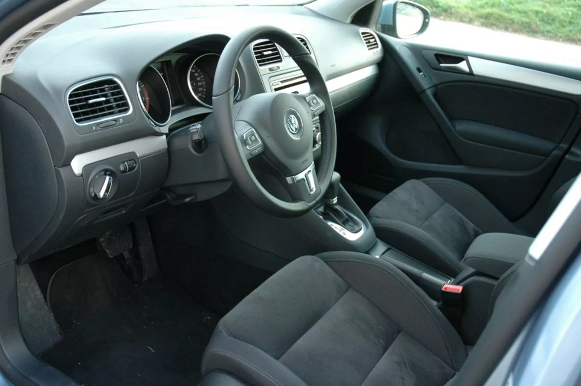 Interiér VW Golf 2008 zobrazující stranu řidiče z pohledu zadního cestujícího s volantem, palubní deskou, středovou konzolou, pákou automatické převodovky, sedadly potaženými tmavou látkou, ovládacími prvky na dveřích řidiče, kobercovou podlahou s rohožemi a přirozeným osvětlením