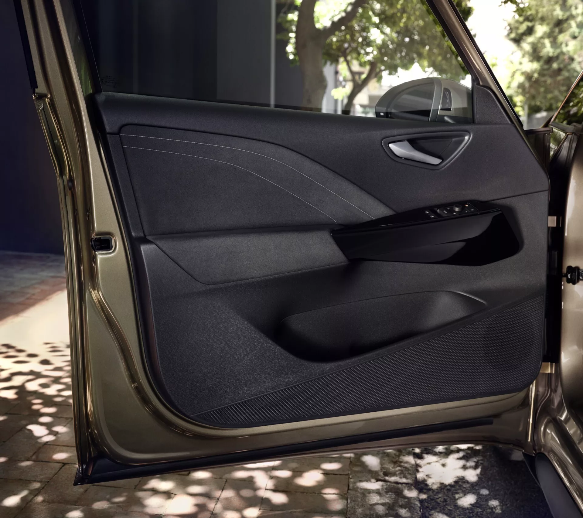Detailní pohled na interiér předních dveří řidiče VW ID.3 2023 1. generace facelift, ukazující prémiový vícemateriálový panel dveří se švy, elektronický ovládací panel pro elektrická okna a zrcátka, rukojeť s chromovým povrchem, úložný prostor a vestavěnou mřížku reproduktoru v přirozeném světle se stínováním