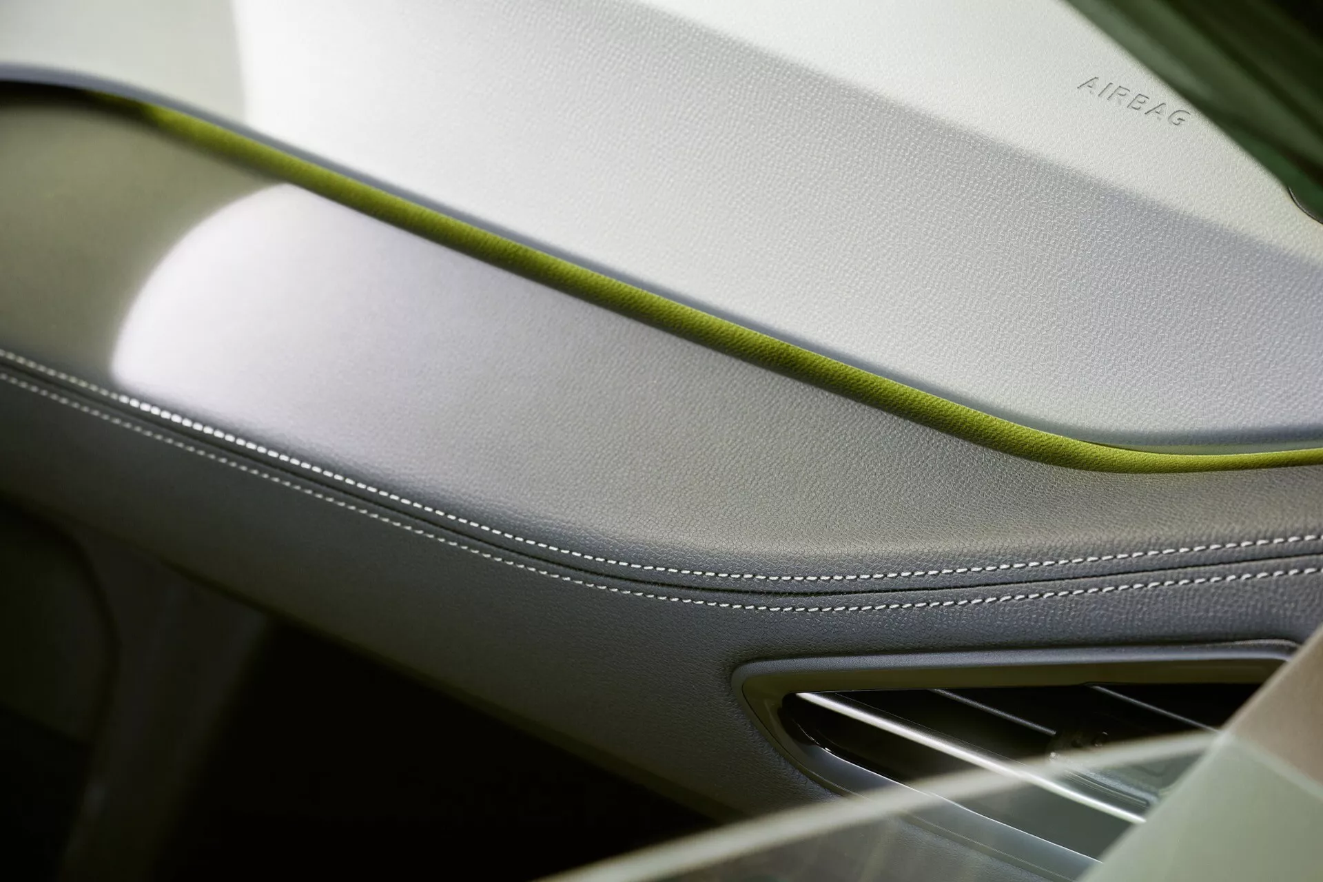 Detailní pohled na interiér VW ID.3 2023 s větrací mřížkou na palubní desce nebo dveřním panelu s krémovým a limetkově zeleným akcentem, zdůrazněním luxusního zpracování, bezpečnostního prvku s vyraženým AIRBAG a chromovým finišem pro pocit vysoké kvality