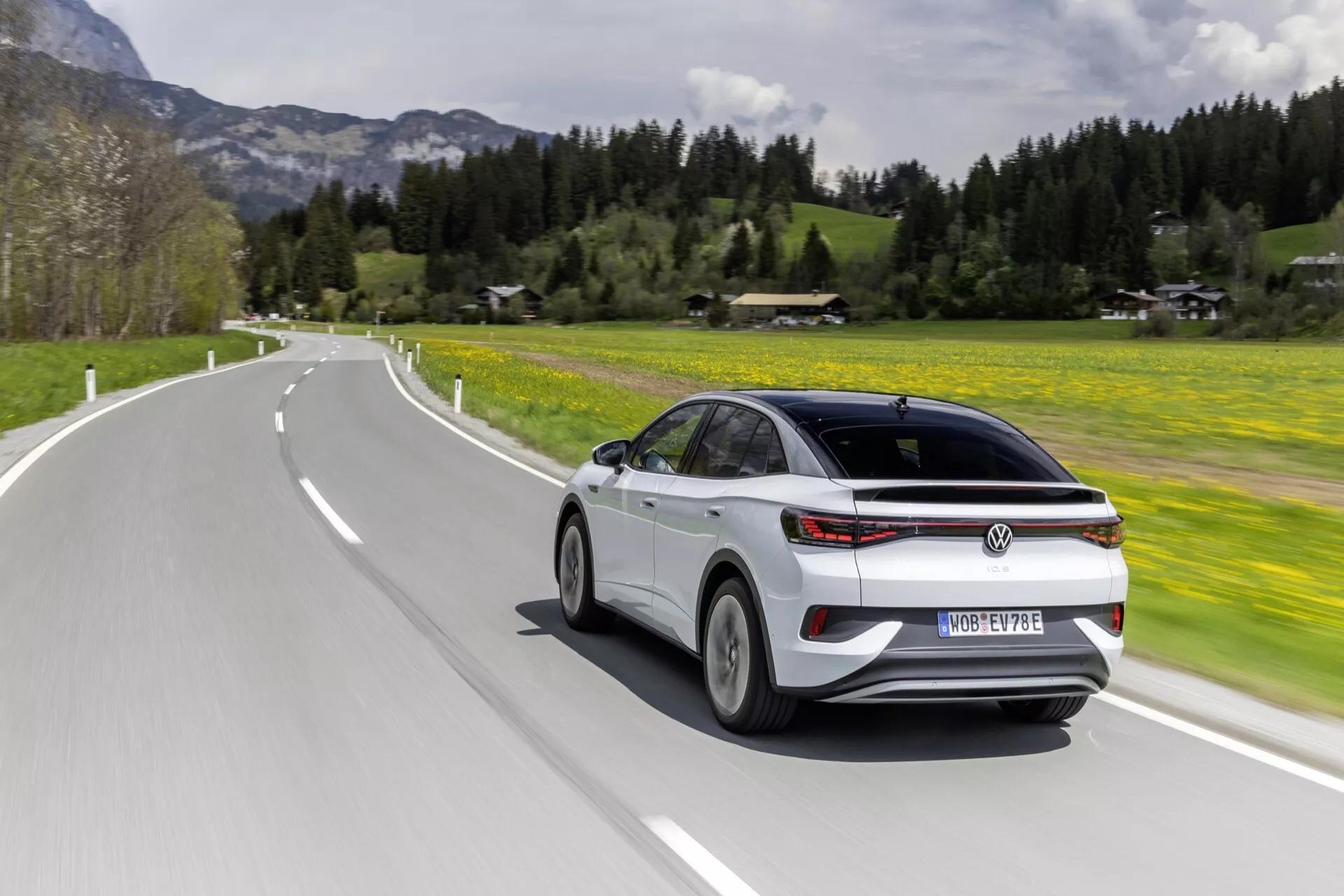 Dynamický pohled na VW ID.5 2022 (1. generace) zezadu tři čtvrtiny při jízdě na silnici. Úhel zdůrazňuje elegantní zadní design vozidla a boční profil, ukazuje zadní prvky jako kufr, zadní okno, zadní světla a zadní nárazník spolu s technologickými prvky jako LED zadní světla a boční zrcátka s integrovanými ukazateli směru na pozadí bujné zelené krajiny.