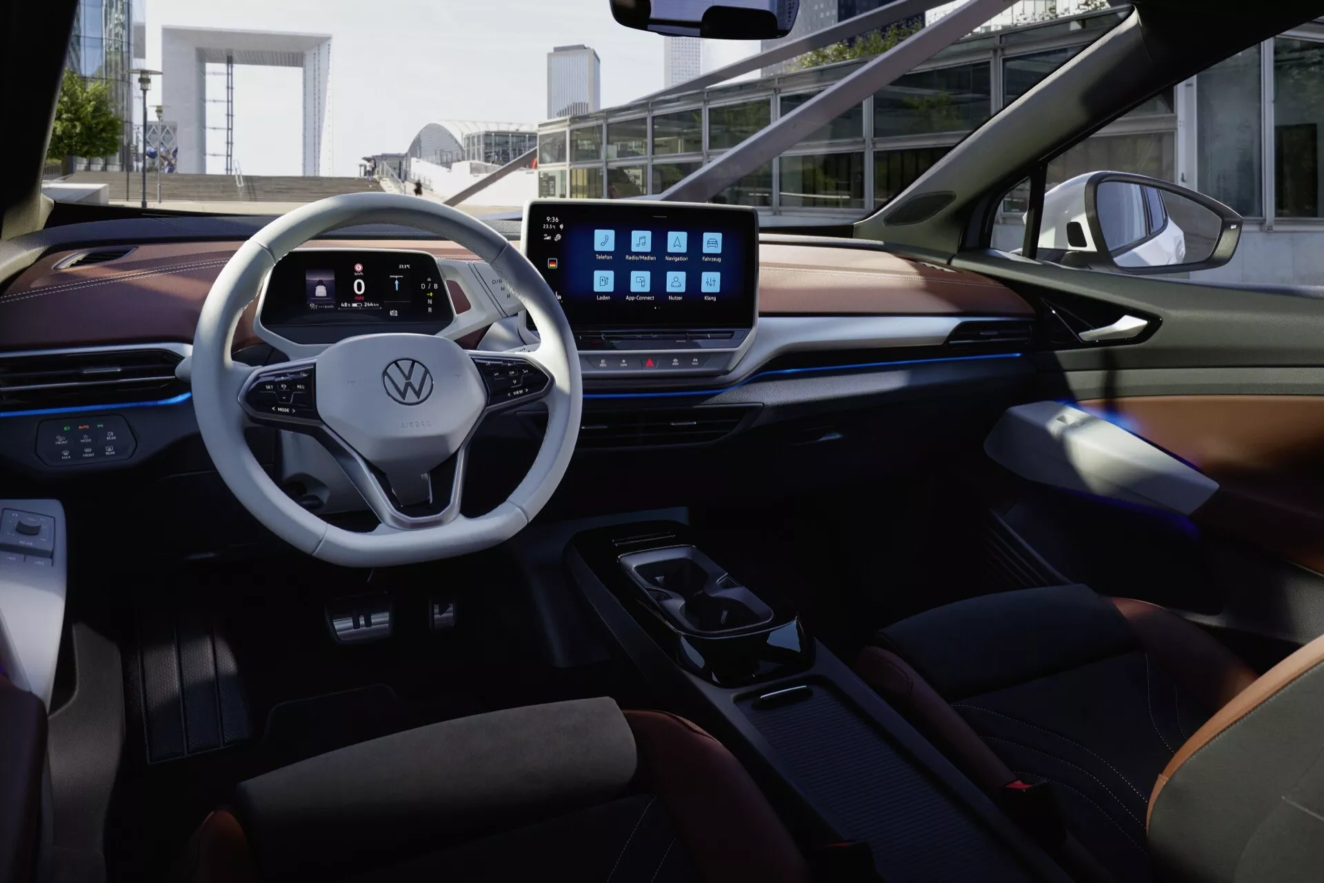 Záběr interiéru modelu VW ID5 2022 (1. generace) z zadního sedadla, zdůrazňující luxus a technologii. Pohled zahrnuje dvouramenný volant s chromovým lemováním, digitální přístrojovou desku, velký dotykový infotainment systém, moderní středovou konzoli a směs kůže a materiálů vzhledu dřeva. Nastavení zdůrazňuje konektivitu s pokročilými funkcemi, jako je Bluetooth a integrace smartphonu, vše vynikající ambientním osvětlením pro útulné prostředí.