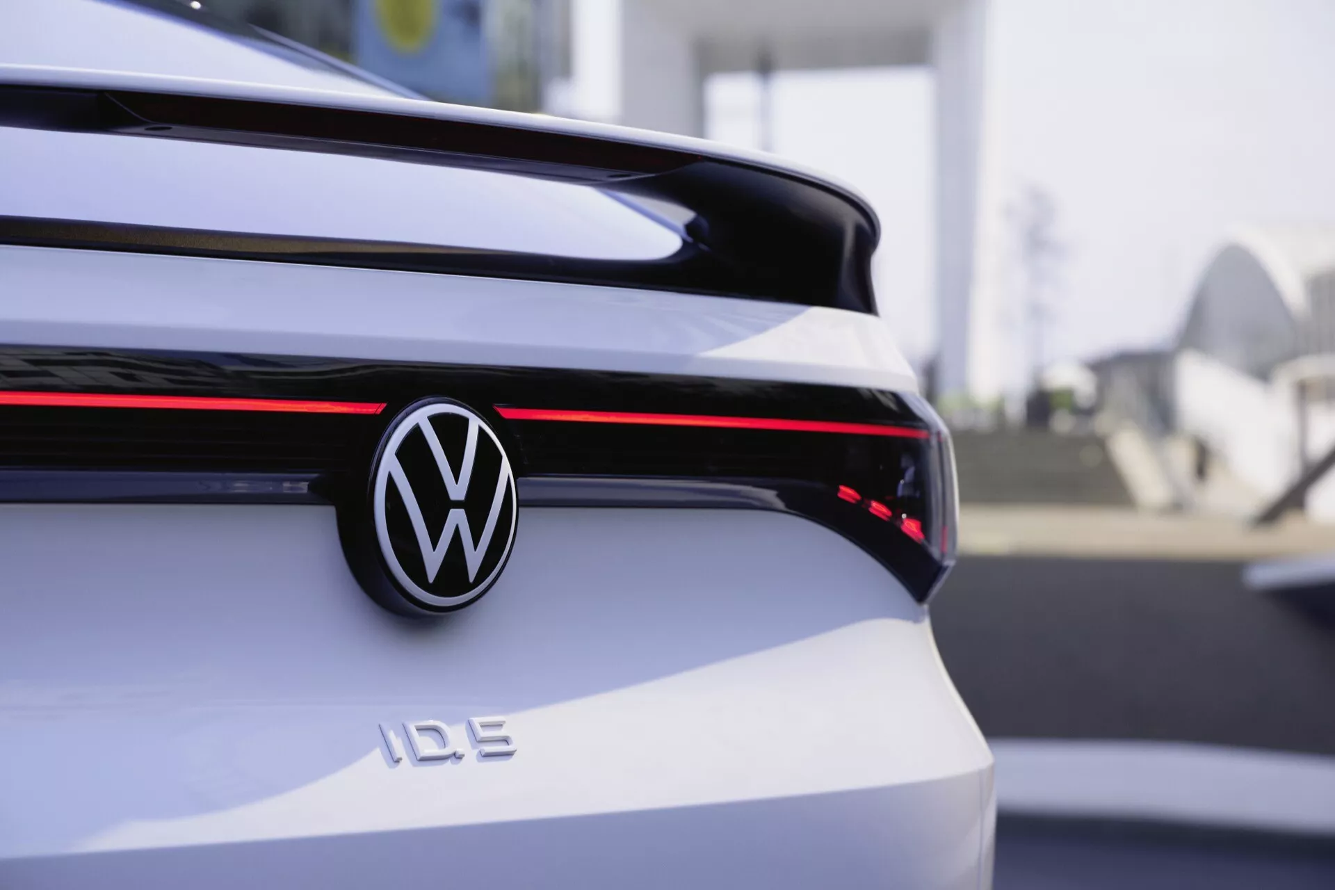 Zadní část elektromobilu Volkswagen ID.5 z roku 2022, představující centrální zavazadlový prostor se zřetelným logem Volkswagen a odznakem 'ID.5'. Horizontální červený světelný pruh se táhne po celé šířce vozu a spojuje zadní světla. Bílý vůz s lesklým povrchem je zobrazen na detailním snímku, mírně mimo střed vlevo, s mělkou hloubkou ostrosti, která rozmazává městské pozadí.