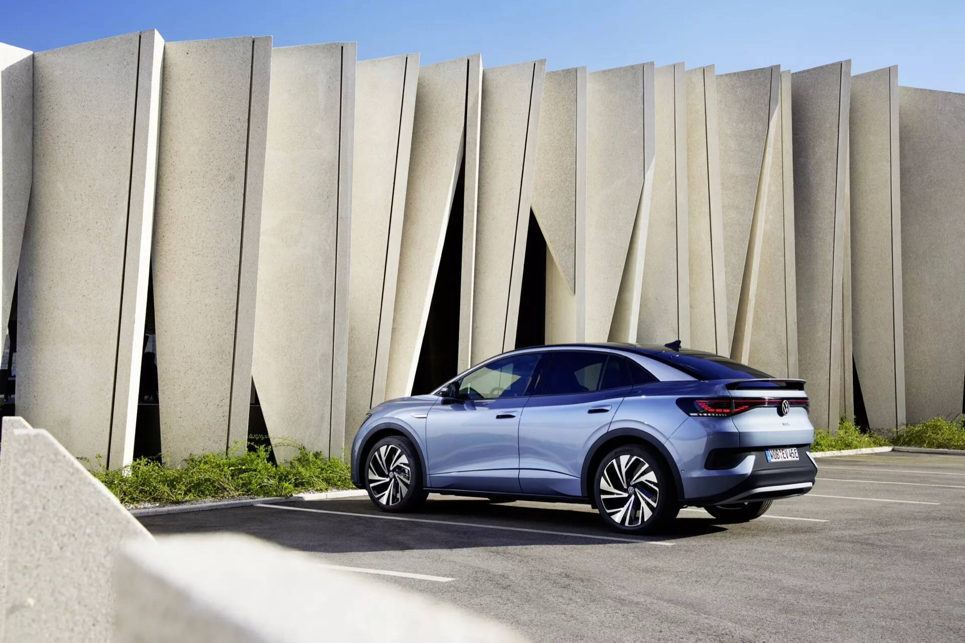 VW ID.5 2022 (1. generace) vyfocený na venkovním místě s moderním architektonickým pozadím. Pohled ze tří čtvrtin na zadní část vozidla ukazuje jeho elegantní designové prvky, včetně zadního spoileru, LED zadních světel a vícepaprskových slitinových kol. Skloněné zadní sklo a aerodynamické kontury naznačují sportovní design hatchbacku nebo coupe-SUV. Fotografie zachycuje auto během dne a odhaluje jeho moderní technologické funkce a současný vzhled.