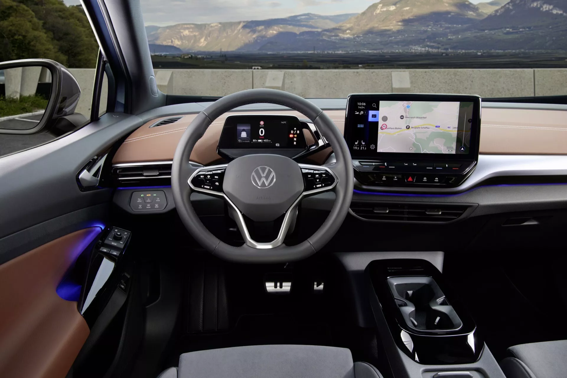 Pohled na interiér moderního Volkswagen ID5 2022 (1. generace) z zadního sedadla. Palubní deska obsahuje velký dotykový infotainment systém, přepínač převodovky a plně digitální přístrojový panel za minimalistickým dvouramenným volantem s dotykovými ovládacími prvky. Interiér kombinuje kůži, měkké plasty a kovové prvky s nastavitelným ambientním osvětlením. Rozložení naznačuje pokročilé propojené vozidlo, pravděpodobně elektrické nebo hybridní.