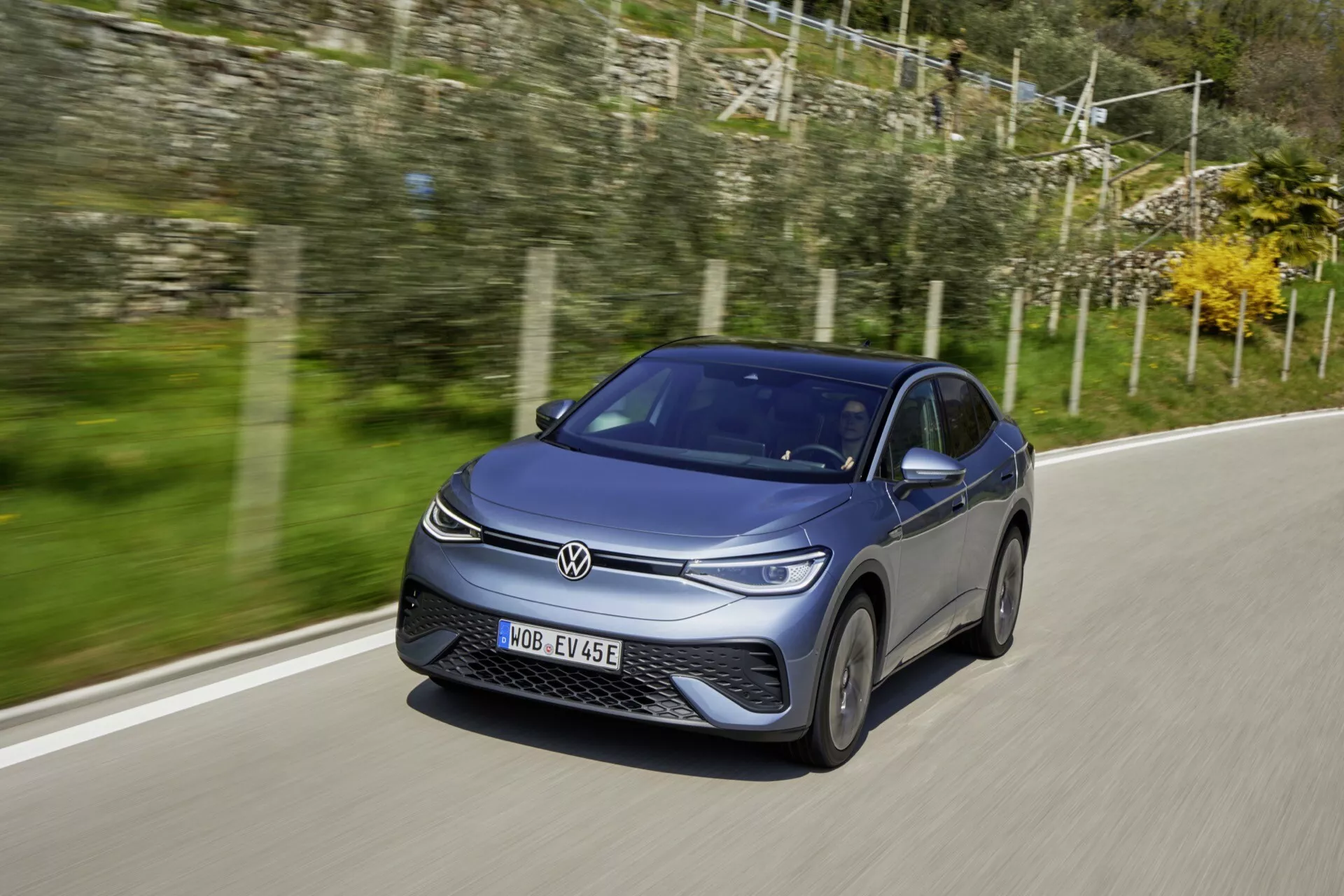 Obrázek zobrazuje moderní hatchback VW ID.5 2022 (1. generace) jedoucí po asfaltové silnici. Fotografován z předního tříčtvrtečního pohledu, zdůrazňuje přední část a stranu řidiče s jasným pohledem na mřížku, světlomety s LED denními světly a v pohybu zachycená litá kola. Karoserie má aerodynamické linie a elegantní tmavou barvu nastavenou přirozeným denním světlem. Dynamický postoj auta je zdůrazněn lehce nižším úhlem fotoaparátu a rozmazaným venkovským pozadím, což naznačuje rychlost.