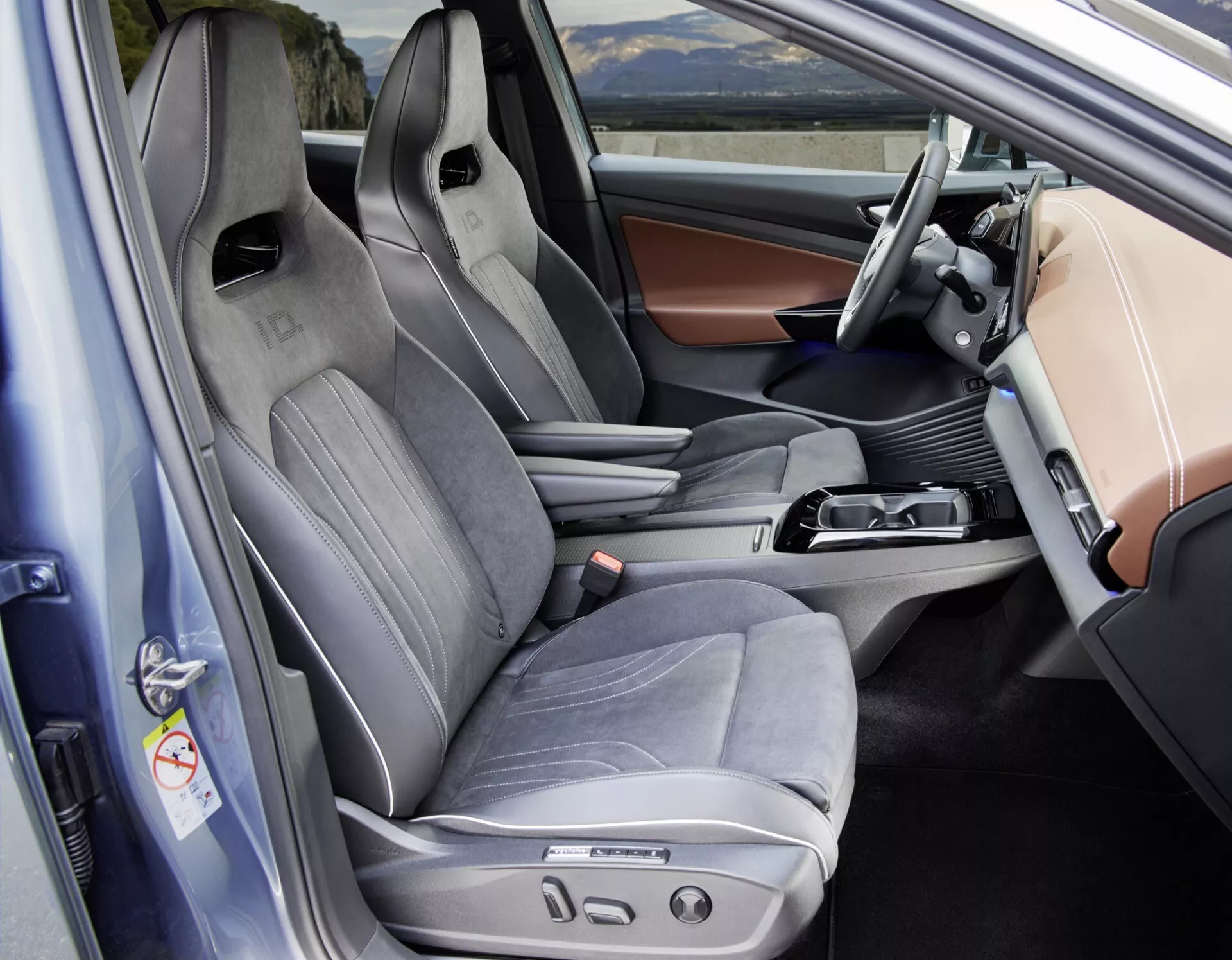 Pohled do interiéru VW ID5 2022 (1. generace) z pohledu řidiče s otevřenými dveřmi, zachycující přední kabinu s důrazem na řidičovo sedadlo a částečně sedadlo spolujezdce. Zdůrazněné prvky zahrnují sportovní řidičovo sedadlo s bočním vedením, kombinaci kůže a textilu a více elektrických ovládacích prvků pro pohodlí. Elegantní centrální konzola obsahuje řadicí páku pro automatickou převodovku, ovládací knoflík pro navigační nebo multimediální systém a malý úložný prostor, což naznačuje použití elektronické parkovací brzdy. Moderní přístrojová deska má displej pro infotainment a záblesk digitálního přístrojového panelu za volantem. Dveřní panel obsahuje ovládací prvky pro elektrické okno a zrcátka, s možnými paměťovými nastaveními pro pozice sedadel. Celý interiér ukazuje pokročilé technologie a prémiové prvky, vhodné pro kategorie jako interiéry aut, automobilové technologie a interiéry luxusních vozidel.