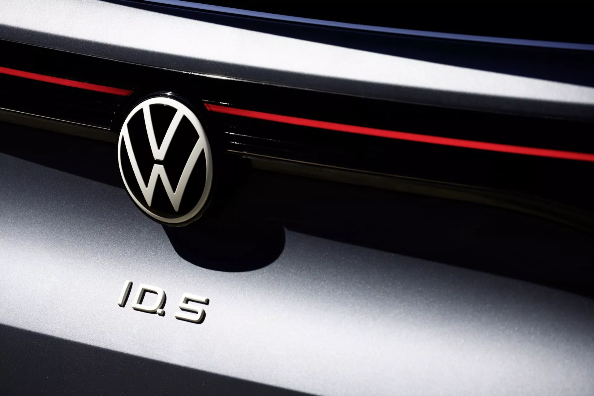 Detailní záběr na Volkswagen ID.5 2022 (1. generace) s ikonickým chromovaným znakem VW na černém kruhovém pozadí a modelovým označením 'ID.5'. Obrázek zdůrazňuje přední část elektrického vozidla se stylovým horizontálním červeným pruhem, který naznačuje sportovní design. Záběr s nízkou hloubkou ostrosti zaměřuje pozornost na detailní loga, přičemž hra stínů a světel zdůrazňuje oblast předního grilu a okraje kapoty.