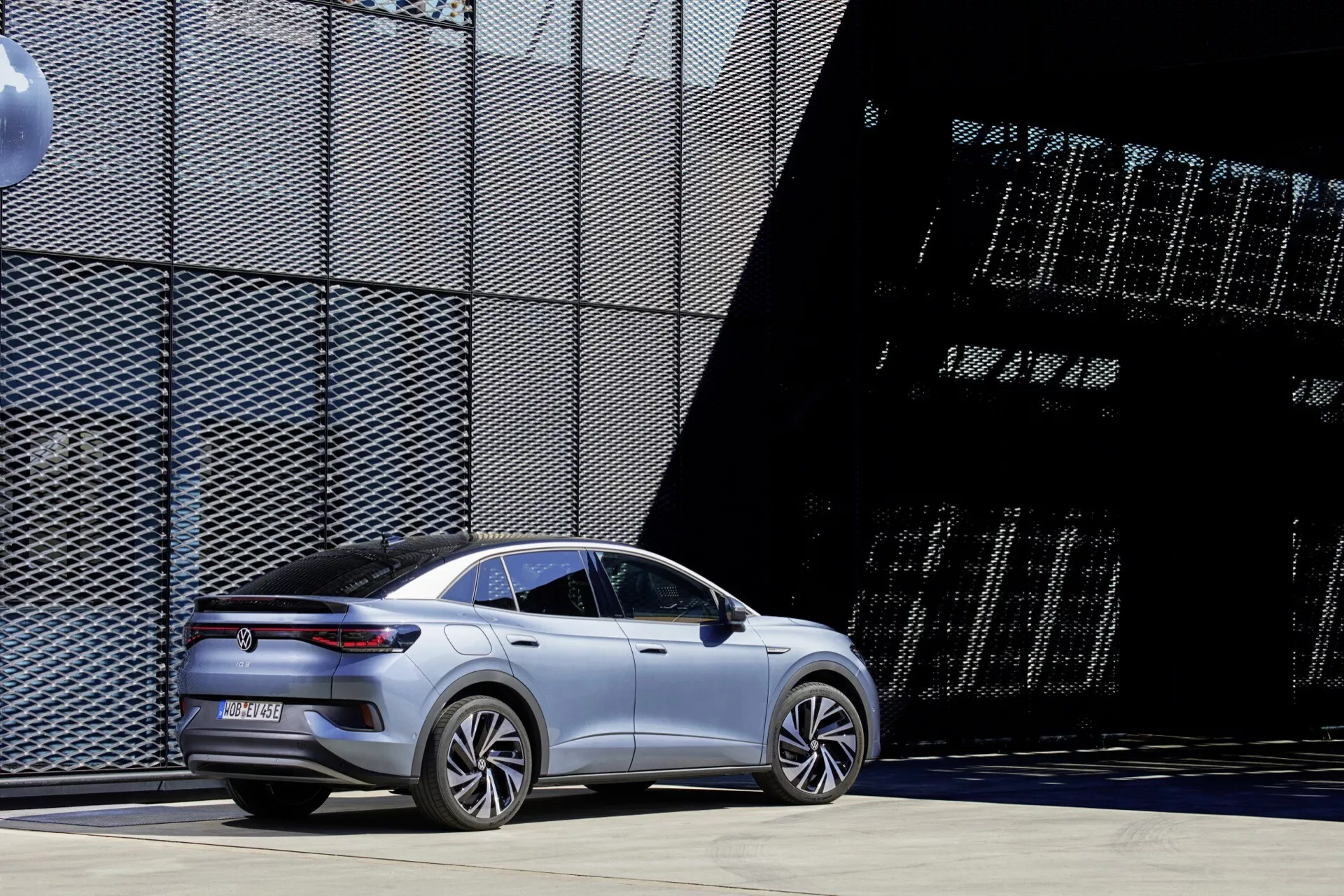 Moderní, stříbrný VW ID.5 2022 první generace sedan se sportovním, aerodynamickým designem zaparkovaný venku během dne se zázemím architektonických struktur v pozadí. Úhel z tří čtvrtin zezadu ukazuje elegantní tělesné linie, zapuštěné kliky dveří, aerodynamická kola, zadní světelnou lištu s LED technologií, velká hliníková kola a svažující se střešní linii, což naznačuje luxusní elektrický nebo hybridní model. Minimalistická estetika bez viditelných výfukových trubek, postavená na zpevněném povrchu s průmyslovým, moderním městským pozadím.