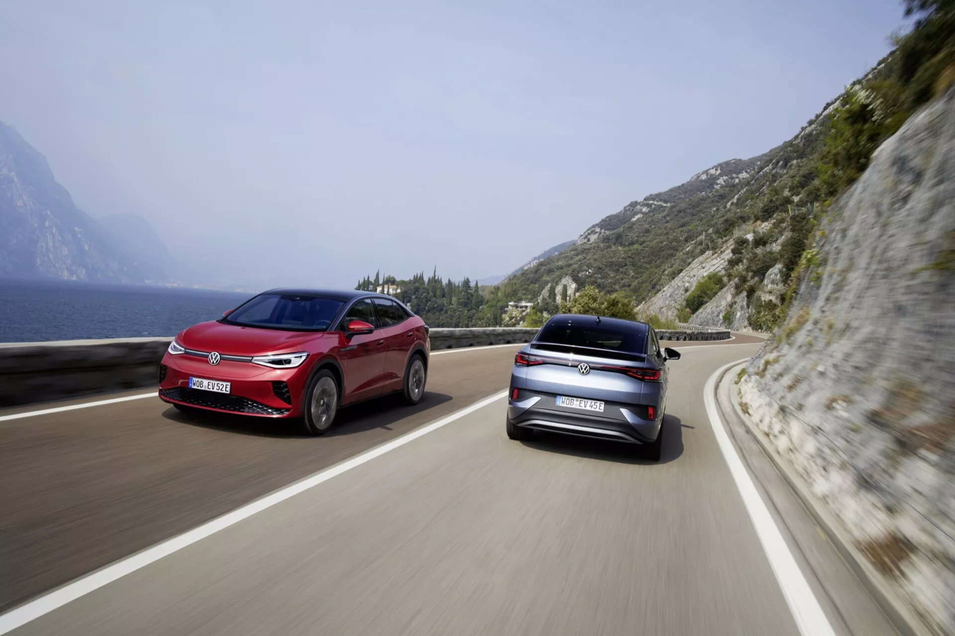 Obrázek ukazuje dvě auta VW ID.5 2022 první generace v pohybu na pobřežní nebo horské silnici pod jasnou oblohou a s kopcovitou, skalnatou krajinou vlevo. Oba vozy jsou zachyceny ze zadního tříčtvrtečního pohledu, což zdůrazňuje moderní, elegantní design s viditelnými zadními světly, kufrem a zadním nárazníkem. Středně velké sedany nebo hatchbacky odrážejí lesklý povrch, což naznačuje vysokou estetickou přitažlivost. Scenérie evokuje pocit cestování a svobody, což naznačuje malebný a dynamický zážitek z jízdy.