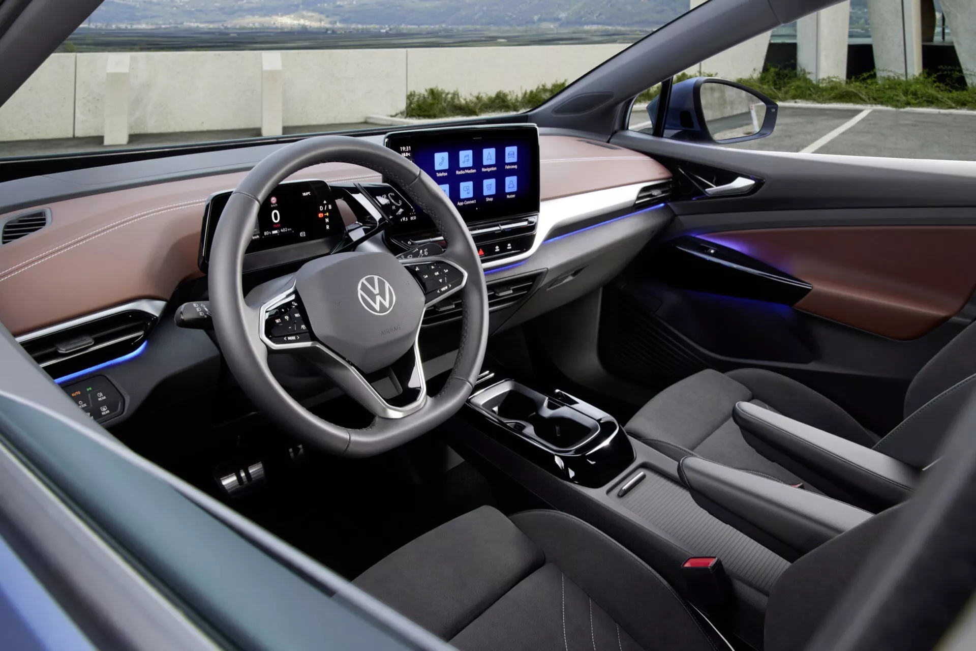 Interiér Volkswagen ID.5 2022 (1. generace) zobrazený z levé strany, zaměřující se na řidičskou oblast a palubní desku. Moderní multifunkční volant s plochým spodkem a logem Volkswagen je obklopen digitálním přístrojovým panelem a velkým dotykovým displejem pro pokročilý infotainment. Palubní deska obsahuje kombinaci materiálů podobných kůži v kontrastních světlých a tmavých tónech. Ambientní osvětlení se táhne přes palubní desku a dveřní panely, zdůrazňuje prémiovou kvalitu a luxusní dokončení interiéru vozidla.